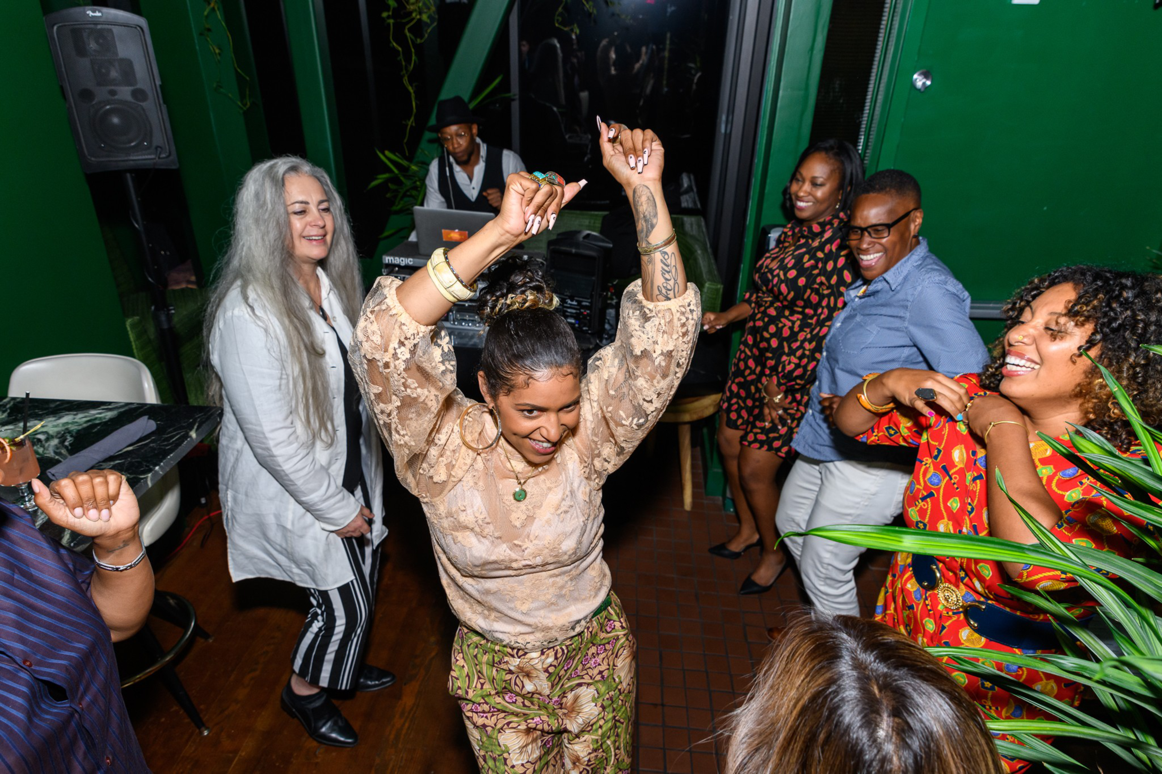 A group of diverse people dance and smile joyfully in a lively room with green walls and wooden floors.