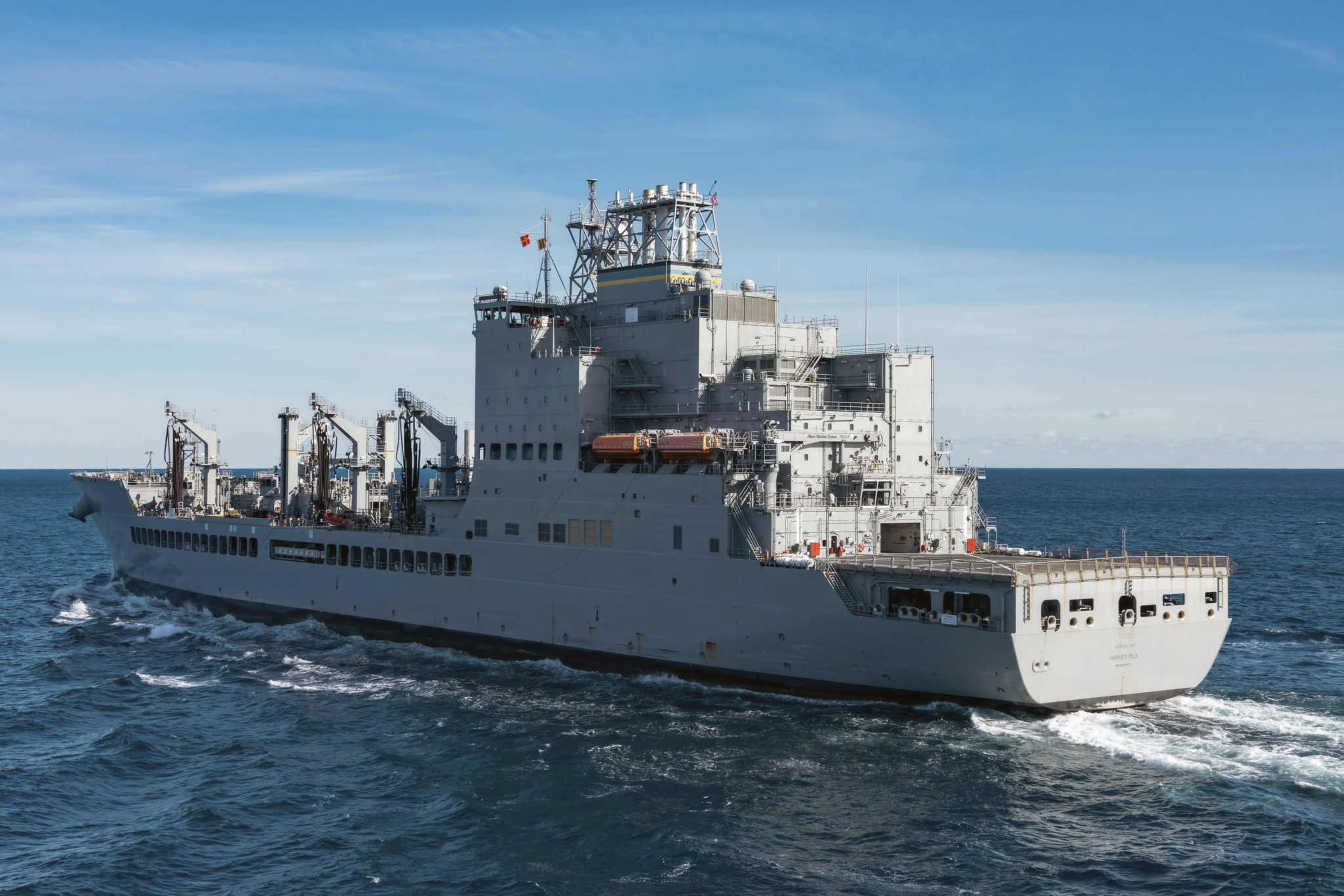 A large gray naval ship sails on the ocean under a clear blue sky. It has multiple decks, cranes, and flags, indicating it's a military support vessel.