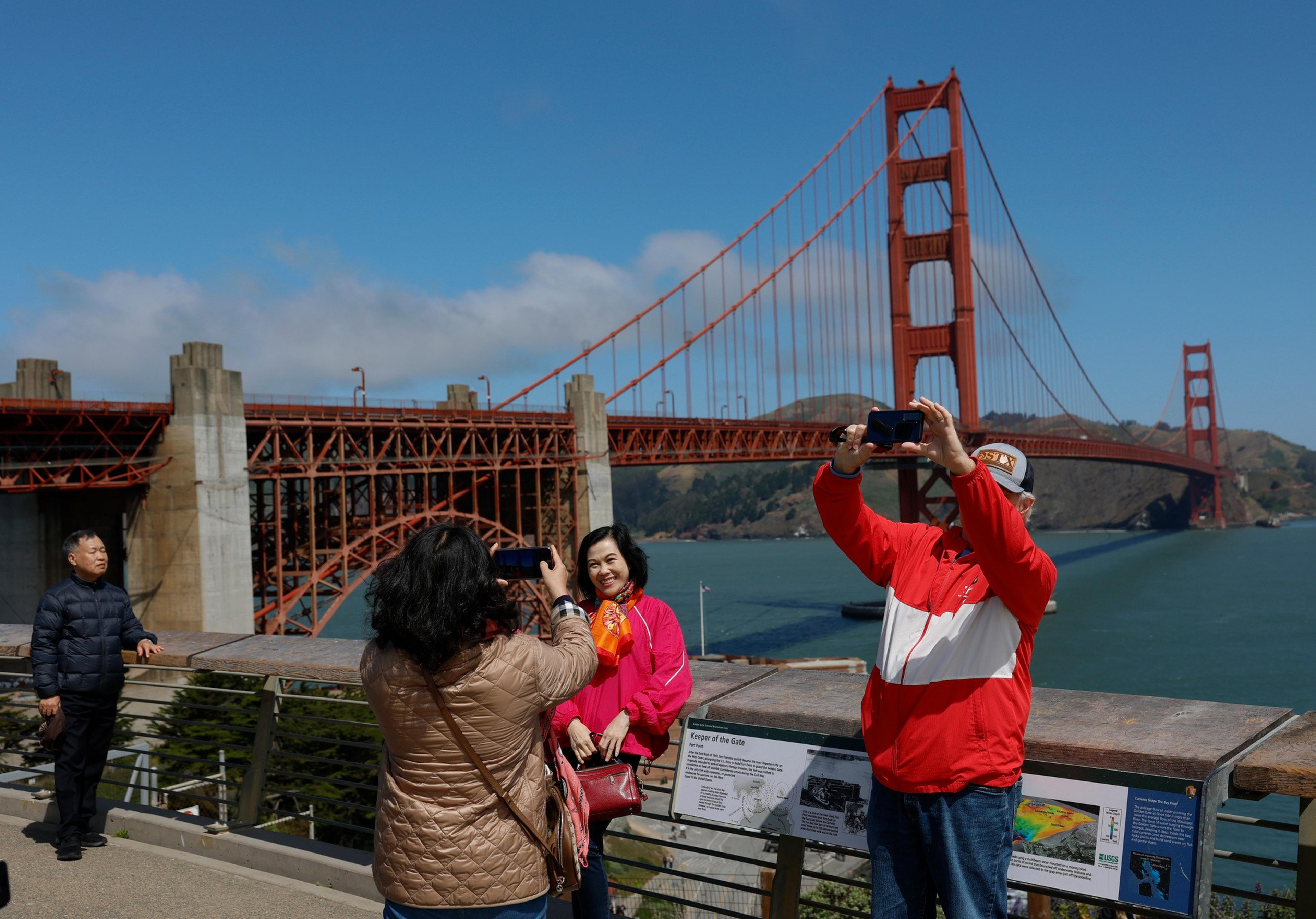 People pose and take selfies near the Golden Gate Bridge on a sunny day with blue skies and calm water in the background.