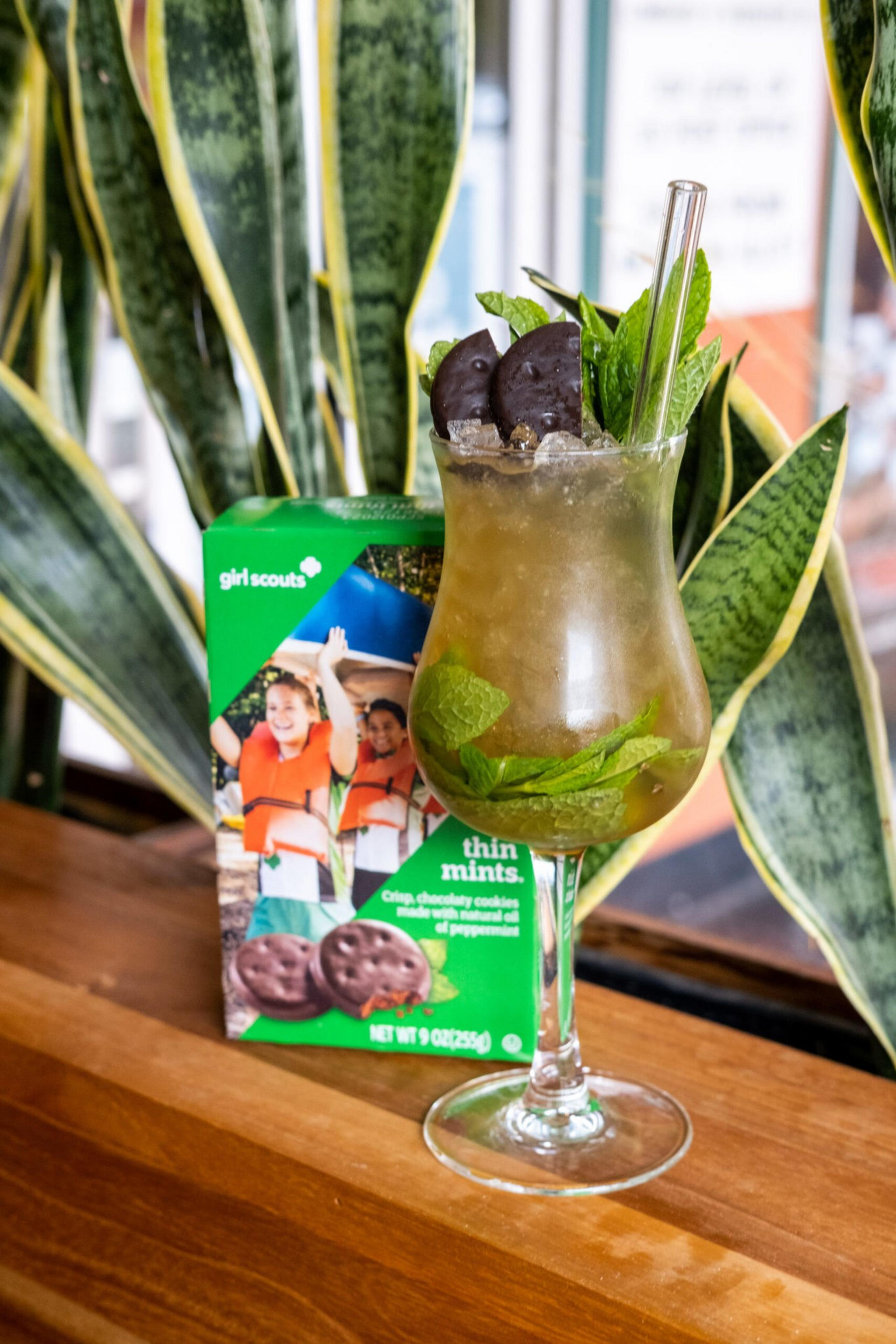 A tall glass with a minty iced drink garnished with Thin Mints cookies and fresh mint leaves sits on a wooden surface beside a box of Girl Scout Thin Mints.