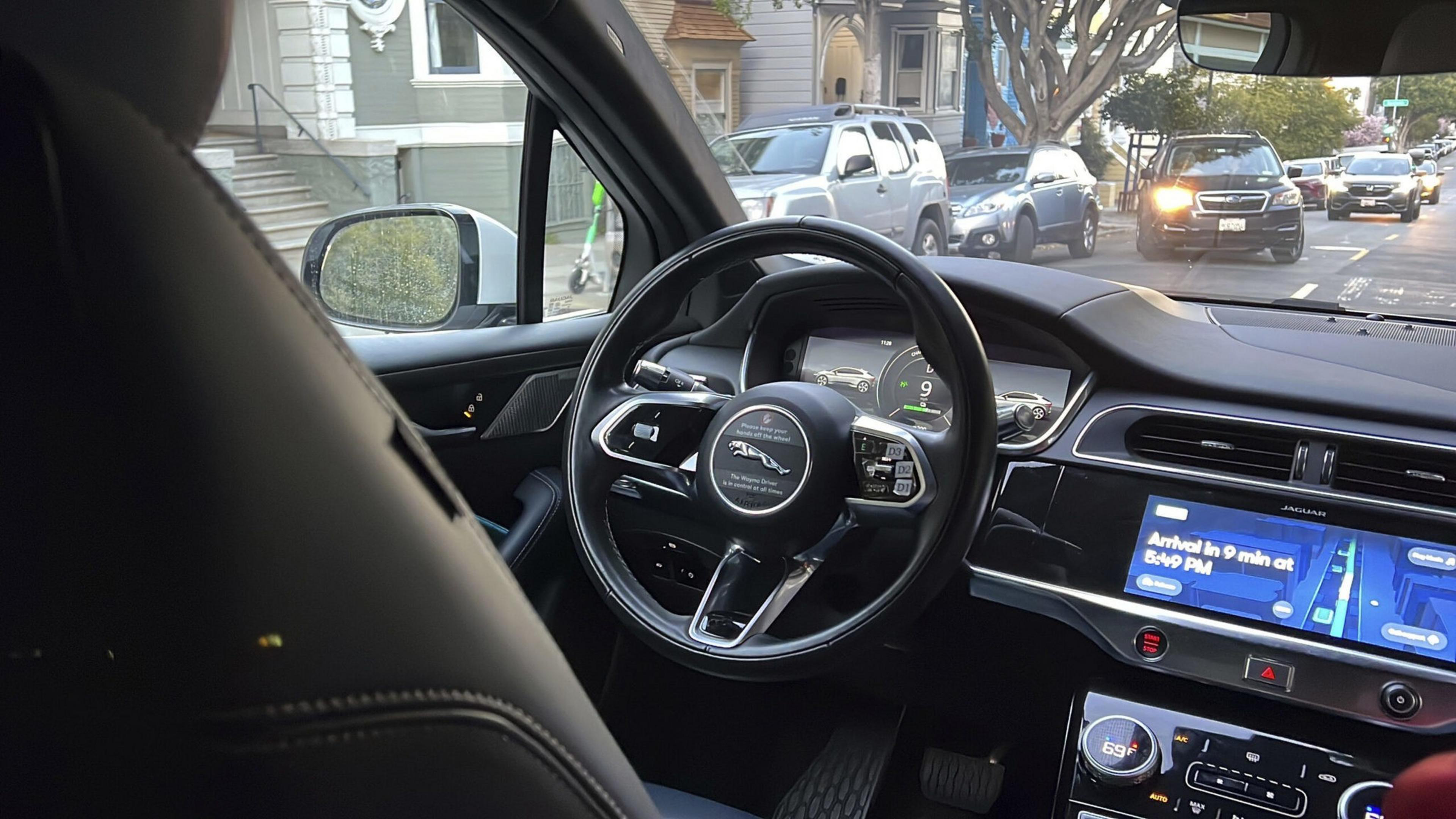 A Jaguar car interior with a digital dashboard, steering wheel, navigation screen, and a street view showing cars and houses outside.
