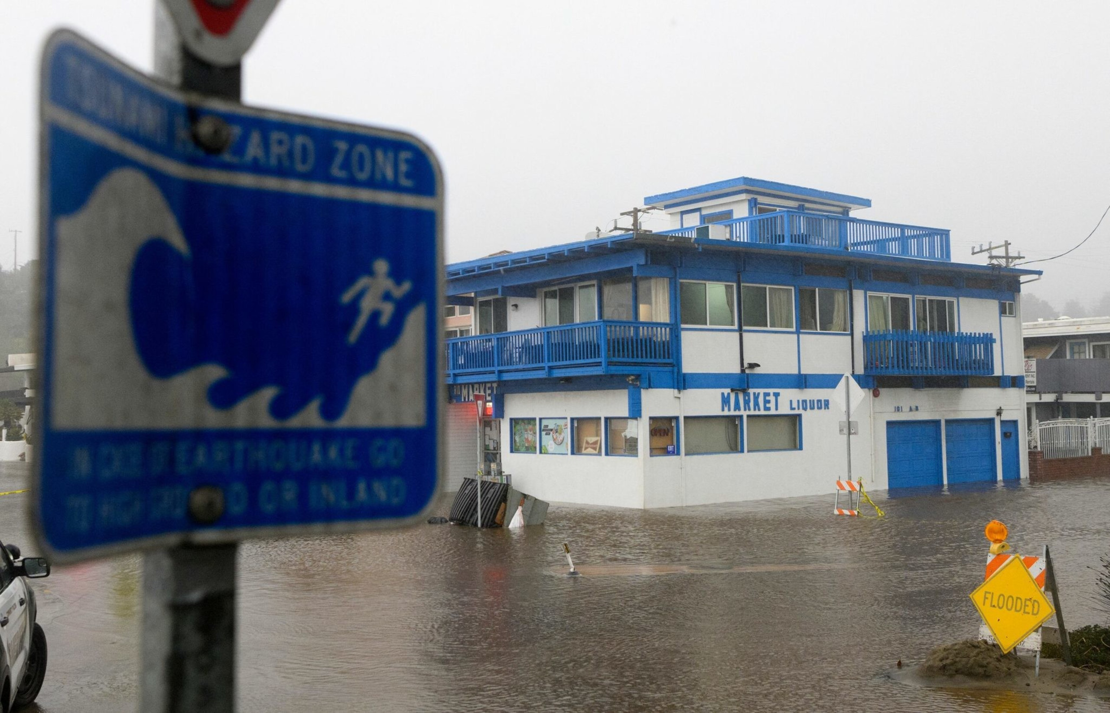 A two-story white and blue building labeled "Market Liquor" is partially submerged in floodwater, with a "Flooded" warning sign and a tsunami hazard zone sign nearby.