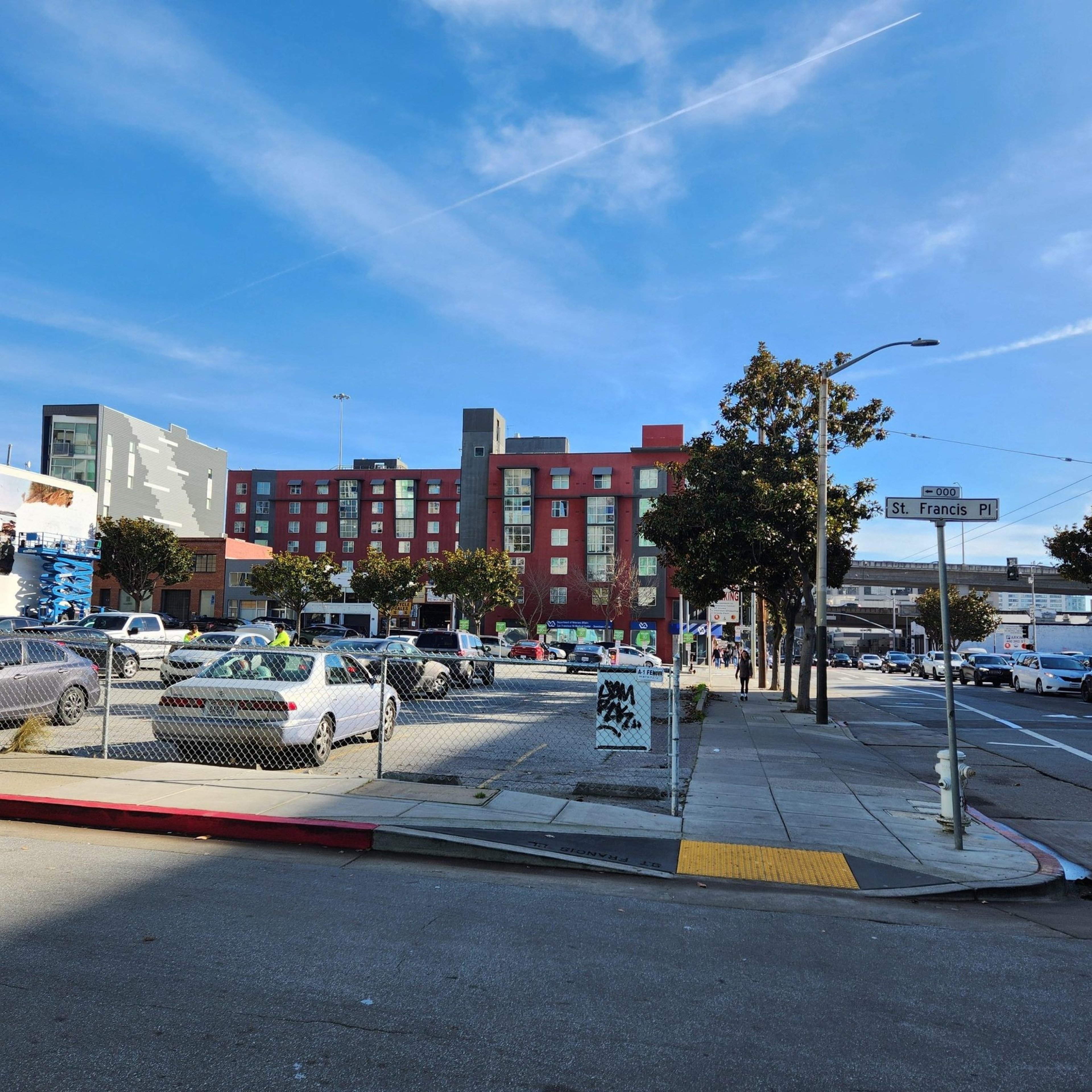 A city street corner with a fenced parking lot, several cars, a red building in the background, trees lining the sidewalk, and a St. Francis Pl street sign.