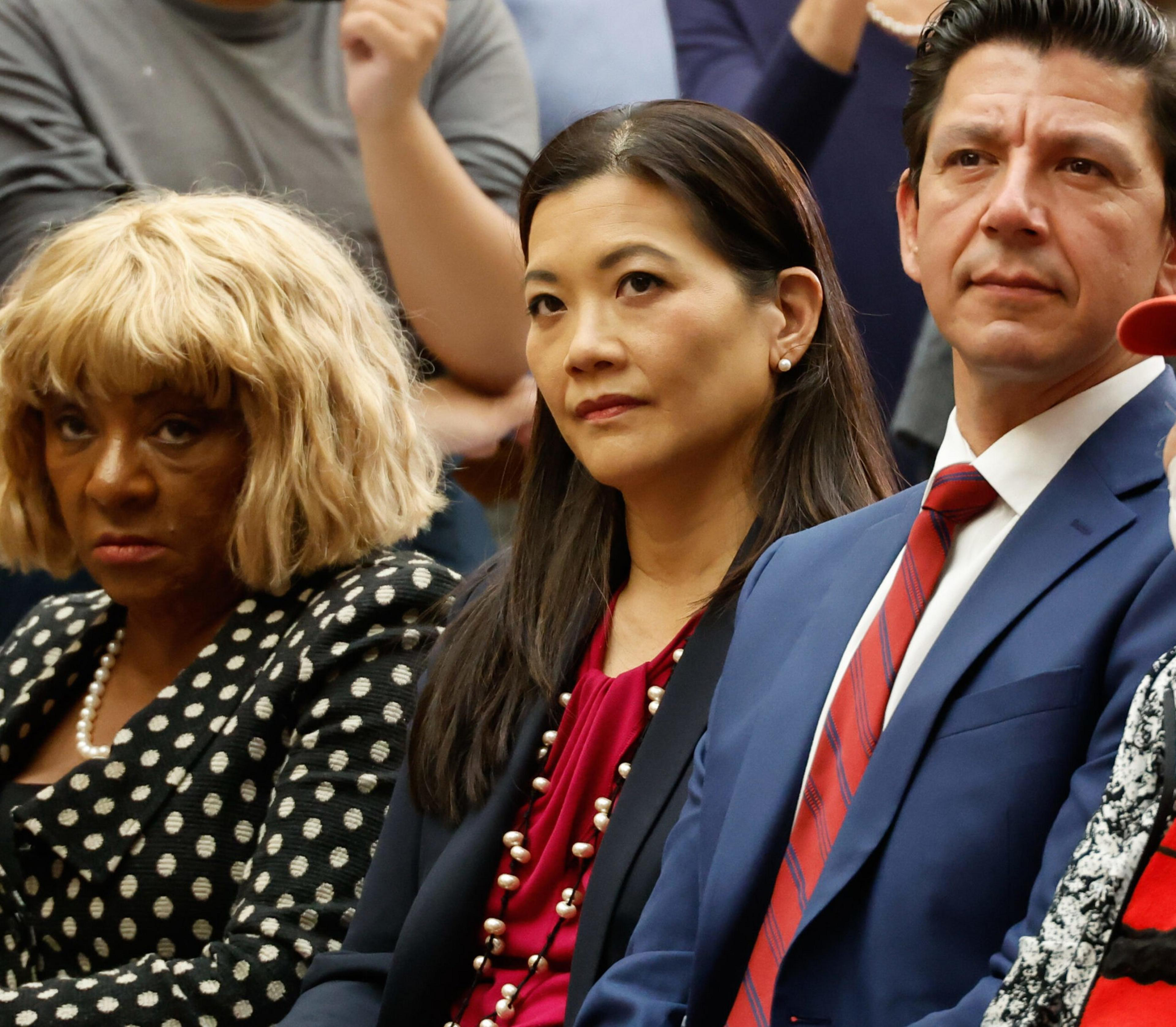 Three adults sit closely together, looking serious; the woman on the left wears a polka-dot outfit, the woman in the middle has a pearl necklace, and the man on the right wears a blue suit with a red striped tie.