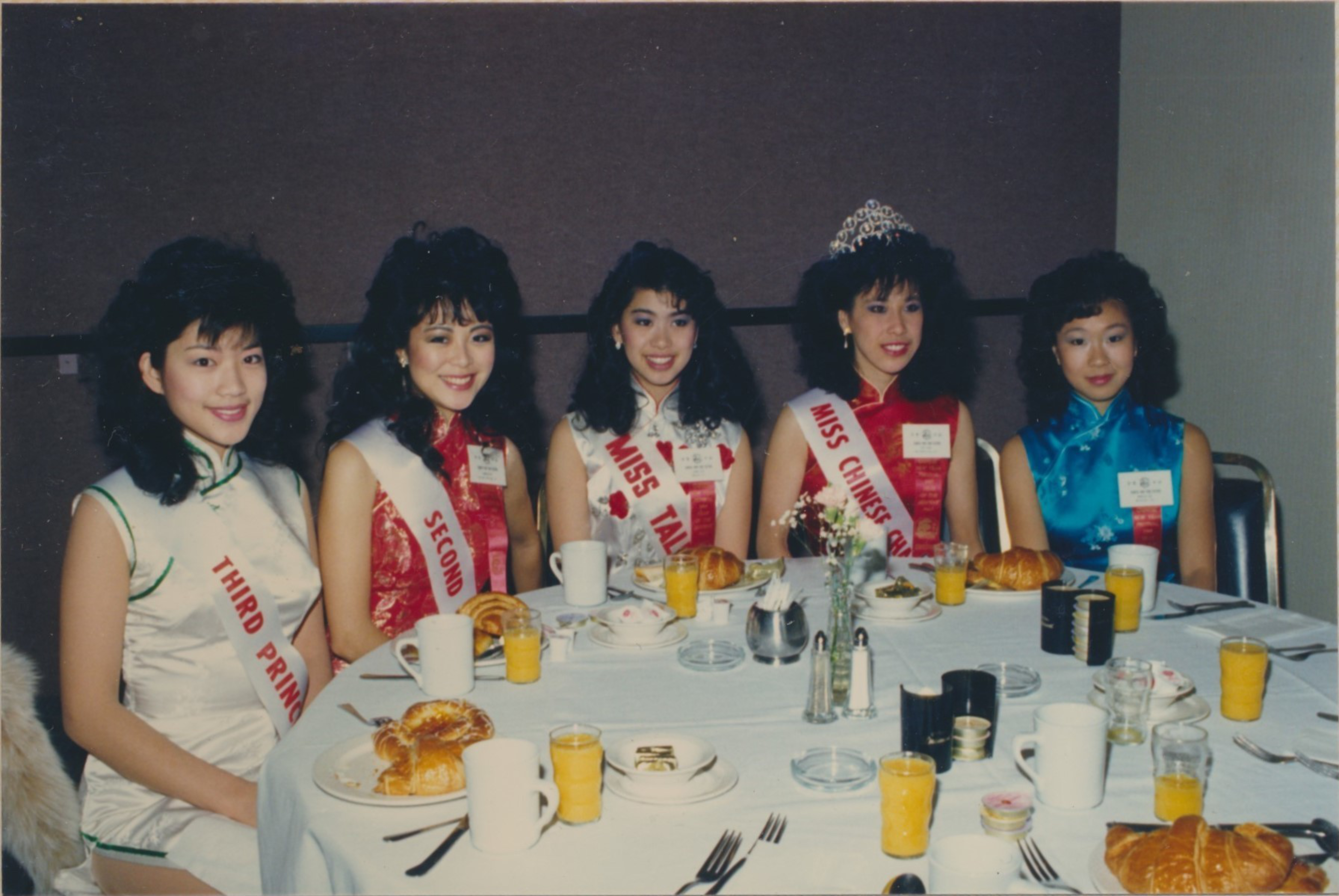 Five women wearing sashes and elegant dresses sit at a round table with breakfast items, including croissants, orange juice, and coffee, in front of them.