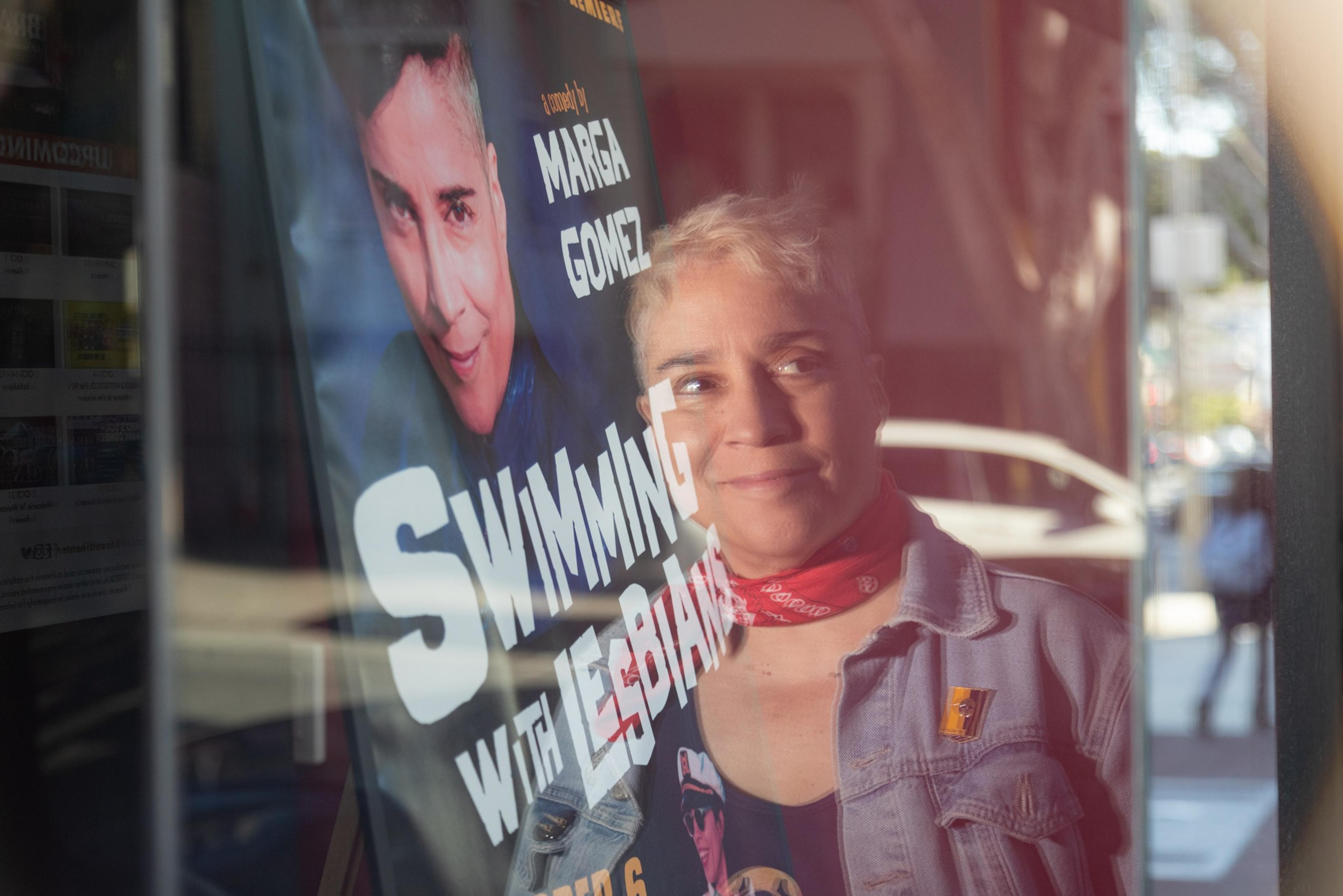 A person with short blonde hair and a red bandana around their neck smiles while their reflection overlaps a poster that reads "Swimming with Lesbians."