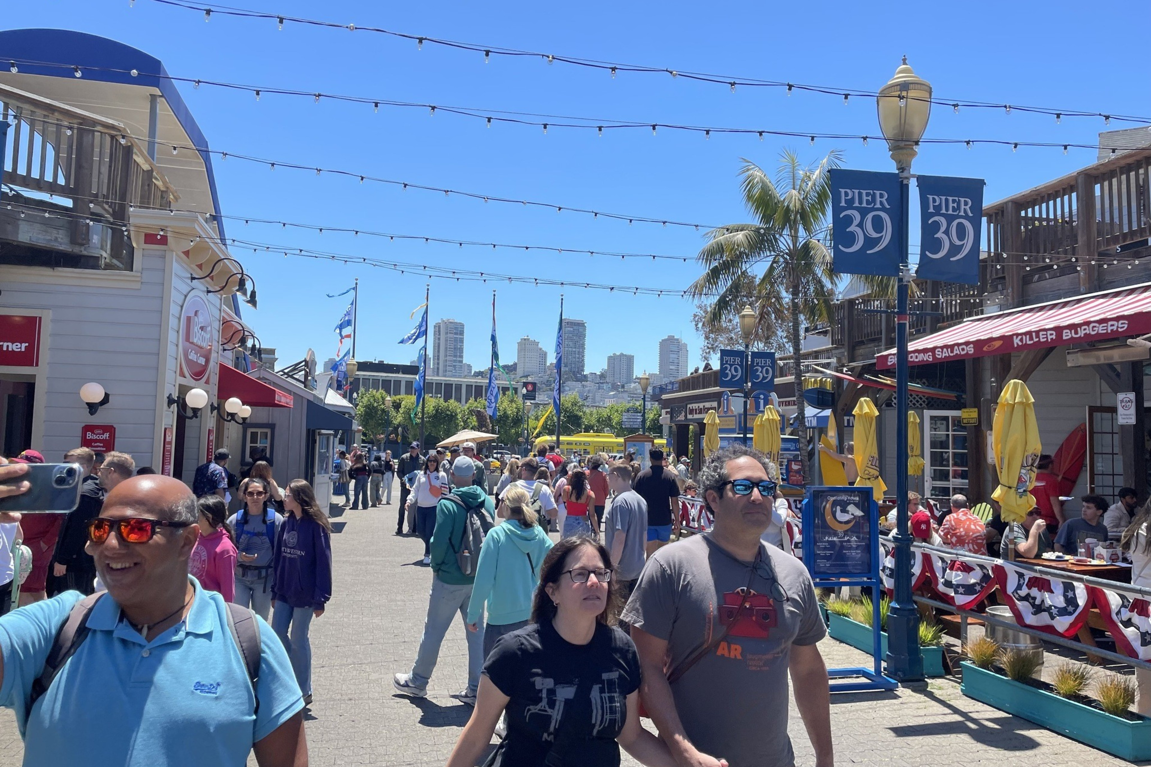 People stroll along a busy, sunlit Pier 39, with colorful storefronts, flags, and a slight urban skyline in the background; one person is taking a selfie.