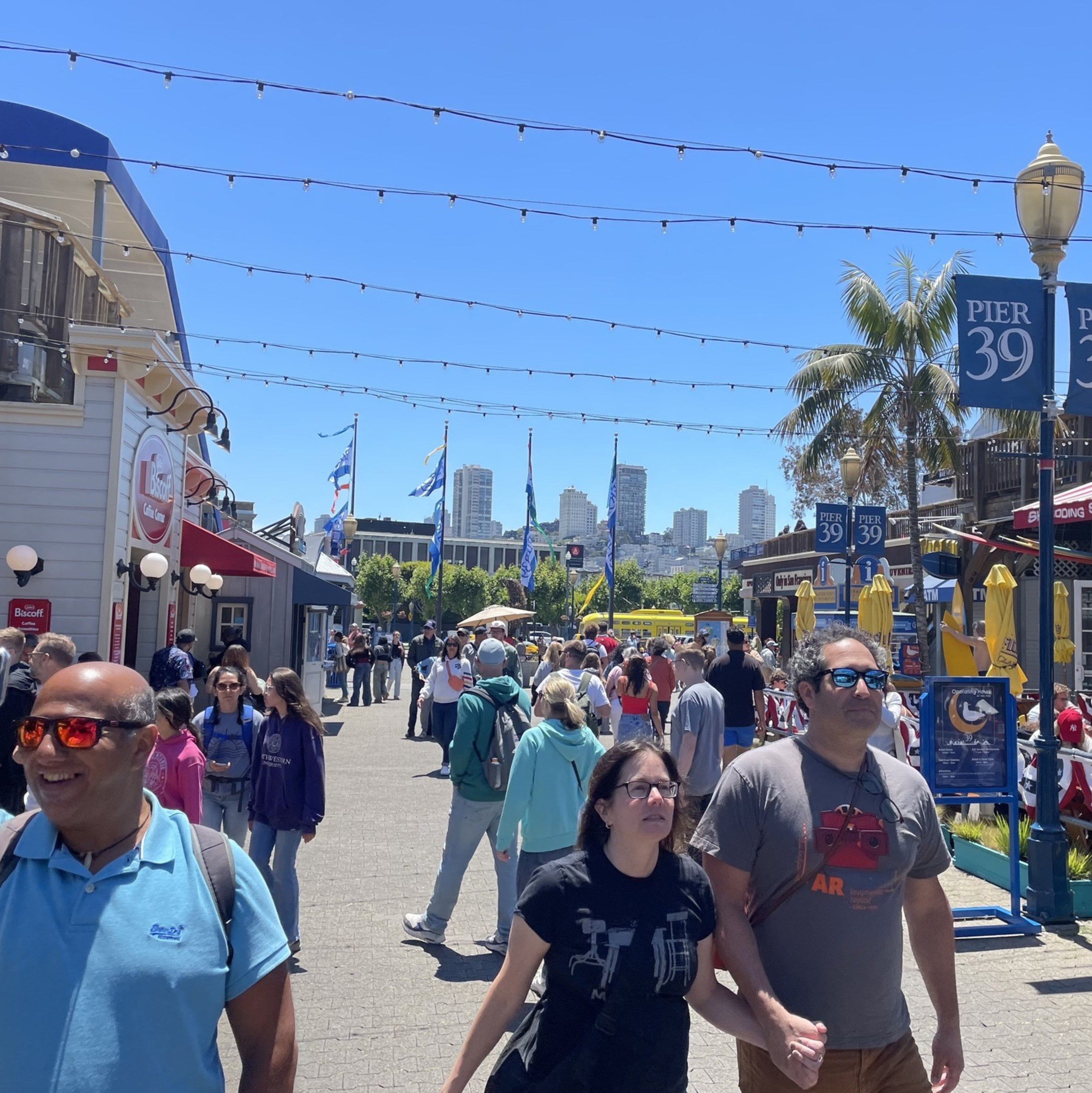 People stroll along a busy, sunlit Pier 39, with colorful storefronts, flags, and a slight urban skyline in the background; one person is taking a selfie.