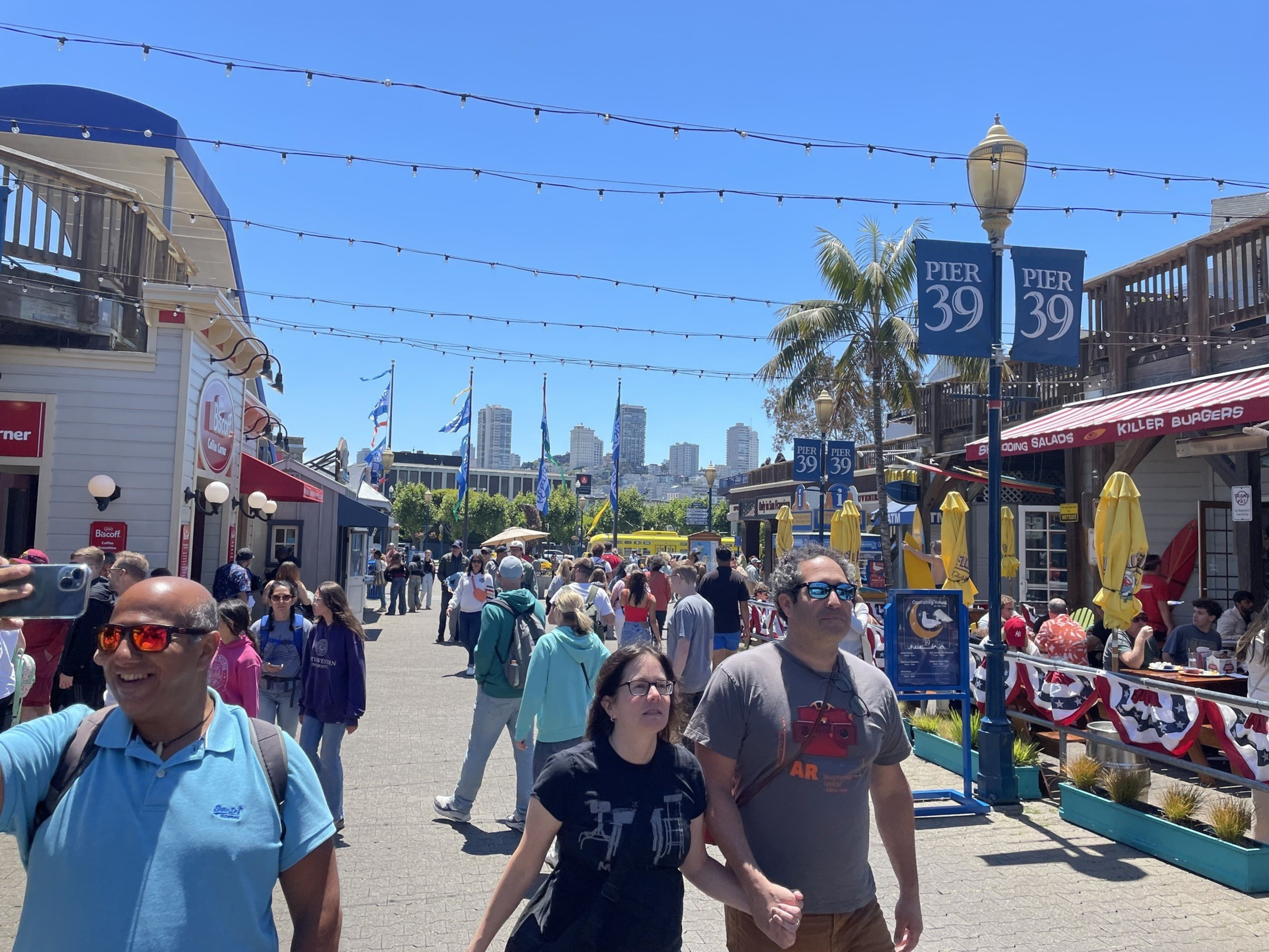 People stroll along a busy, sunlit Pier 39, with colorful storefronts, flags, and a slight urban skyline in the background; one person is taking a selfie.