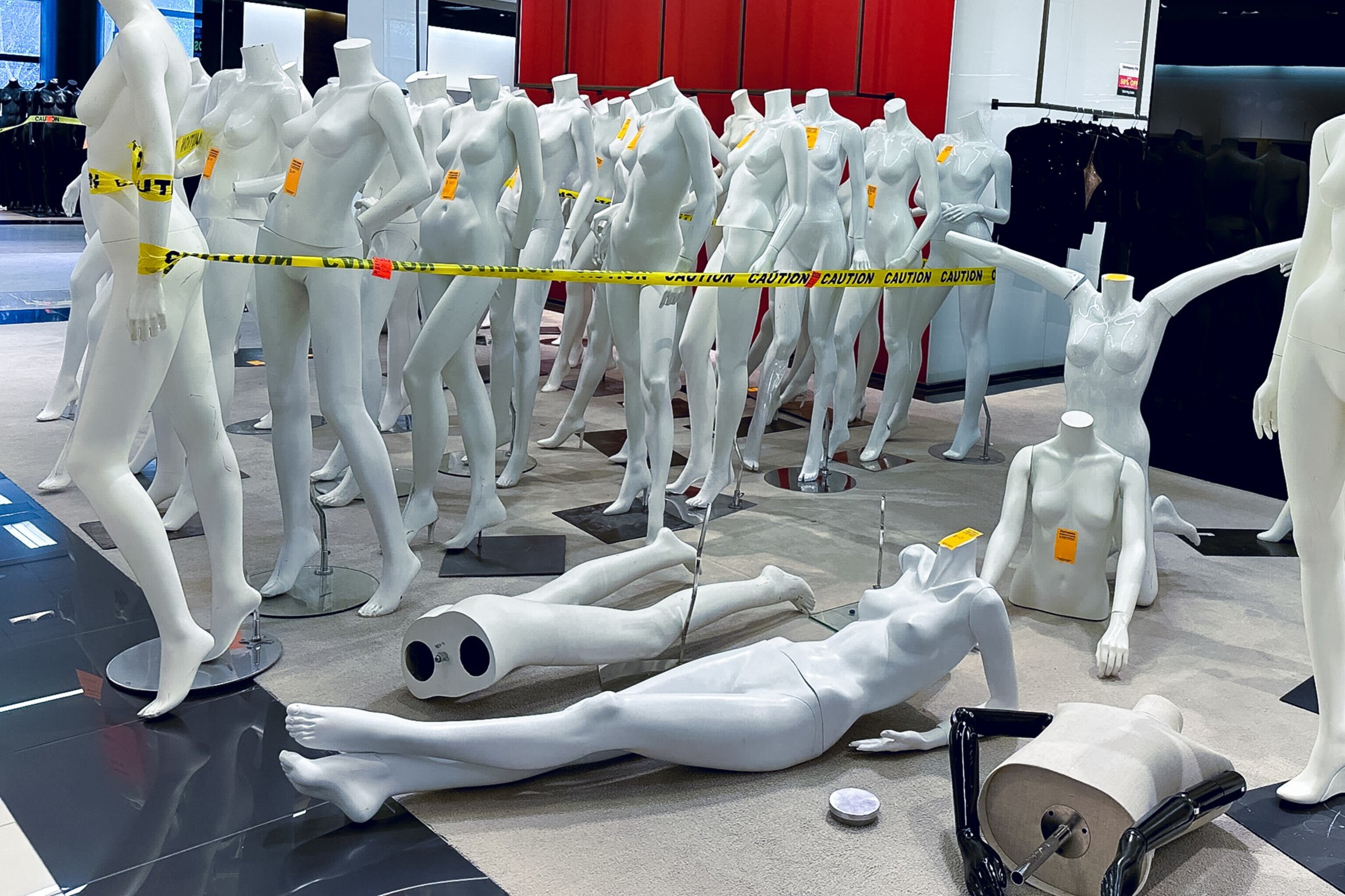A cluttered room with numerous white, headless mannequins and caution tape around them. Some mannequins are standing, while others are scattered on the floor.
