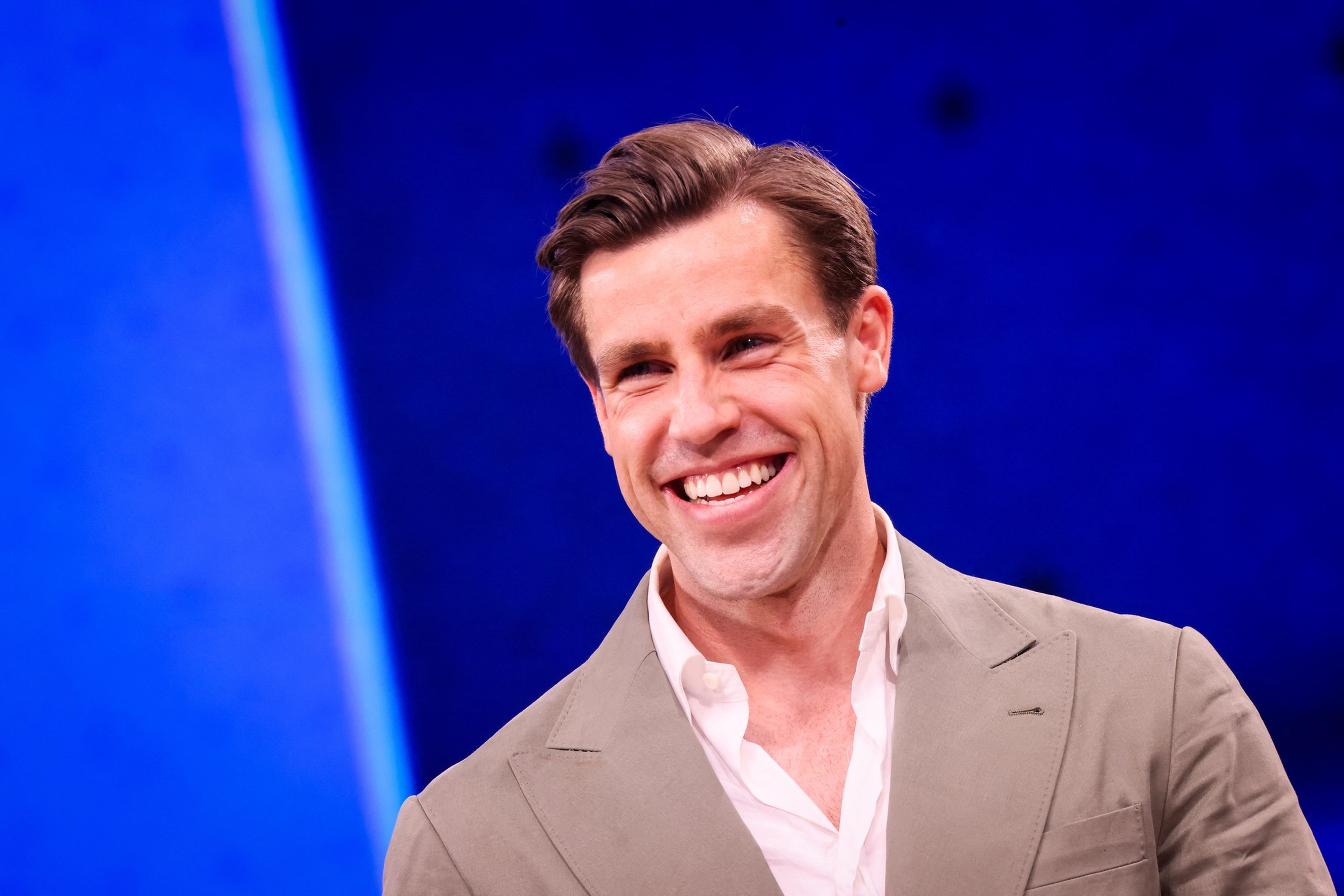 A man with neatly styled brown hair smiles warmly, wearing a light gray suit jacket over an open-collared white shirt against a blue background.