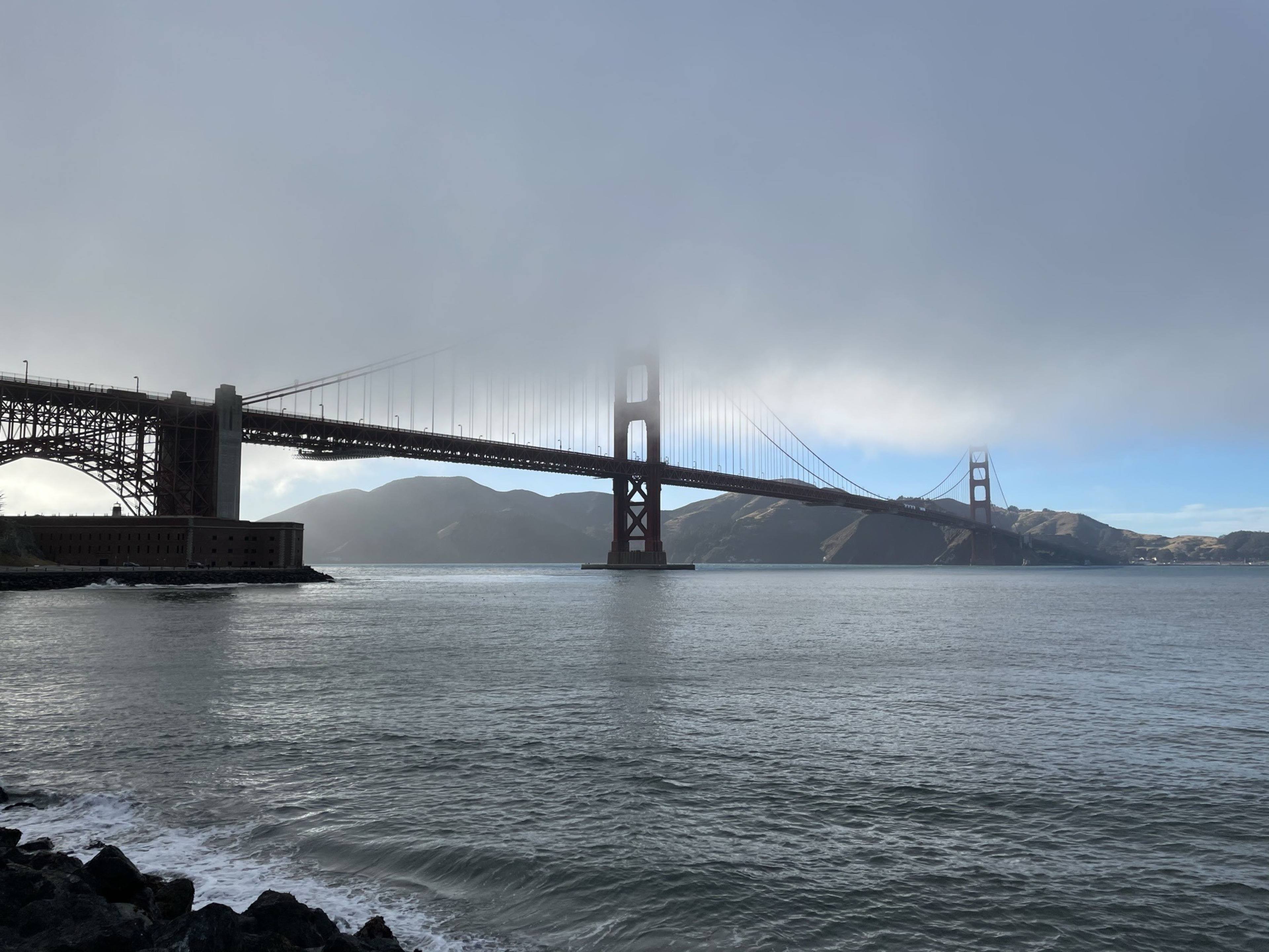 A large suspension bridge spans a calm body of water, partially shrouded in fog. In the background, there are hills under a mostly clear sky.
