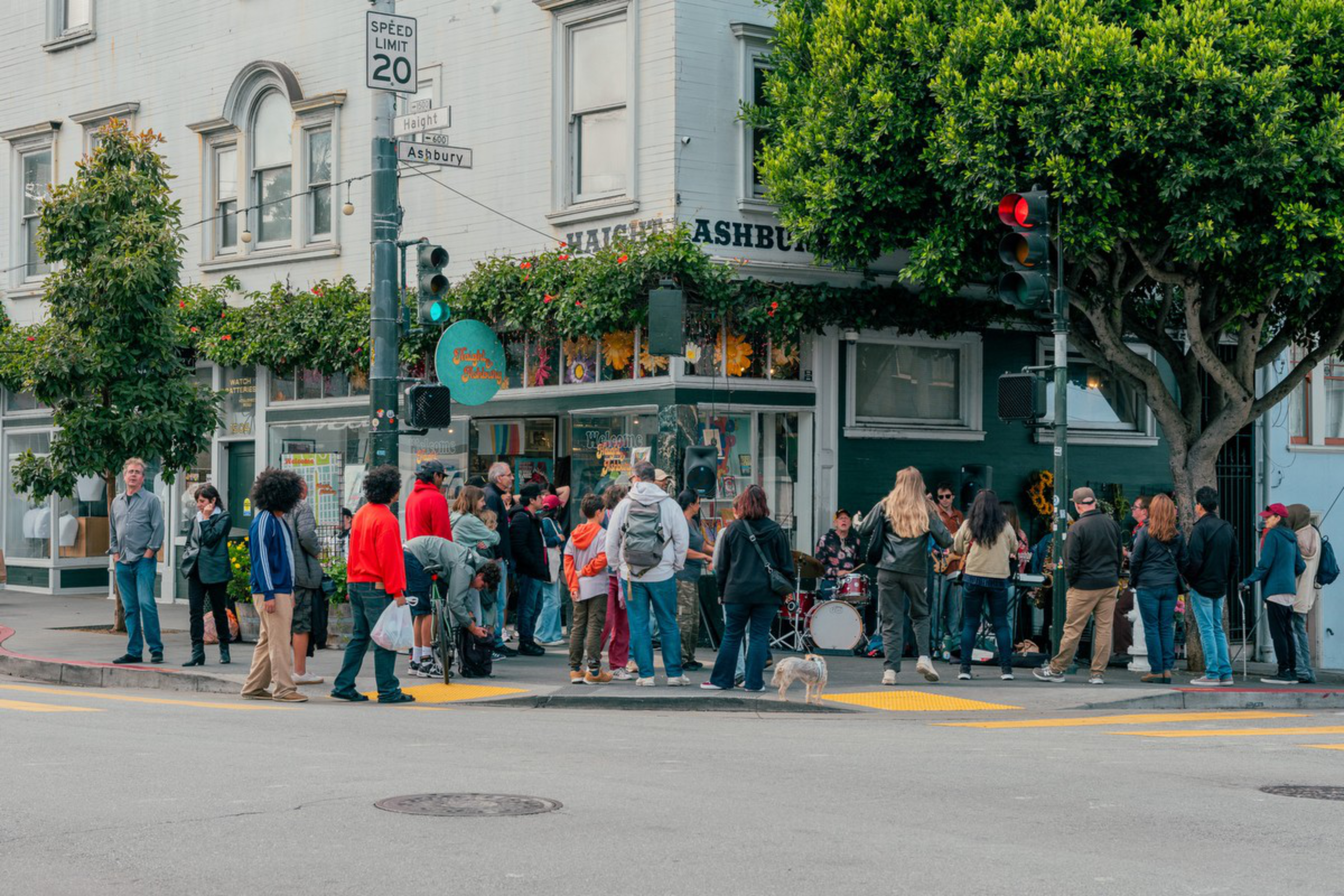 A crowd stands at a corner near a green building with hanging flowers, watching a street band perform by the traffic light and street signs.