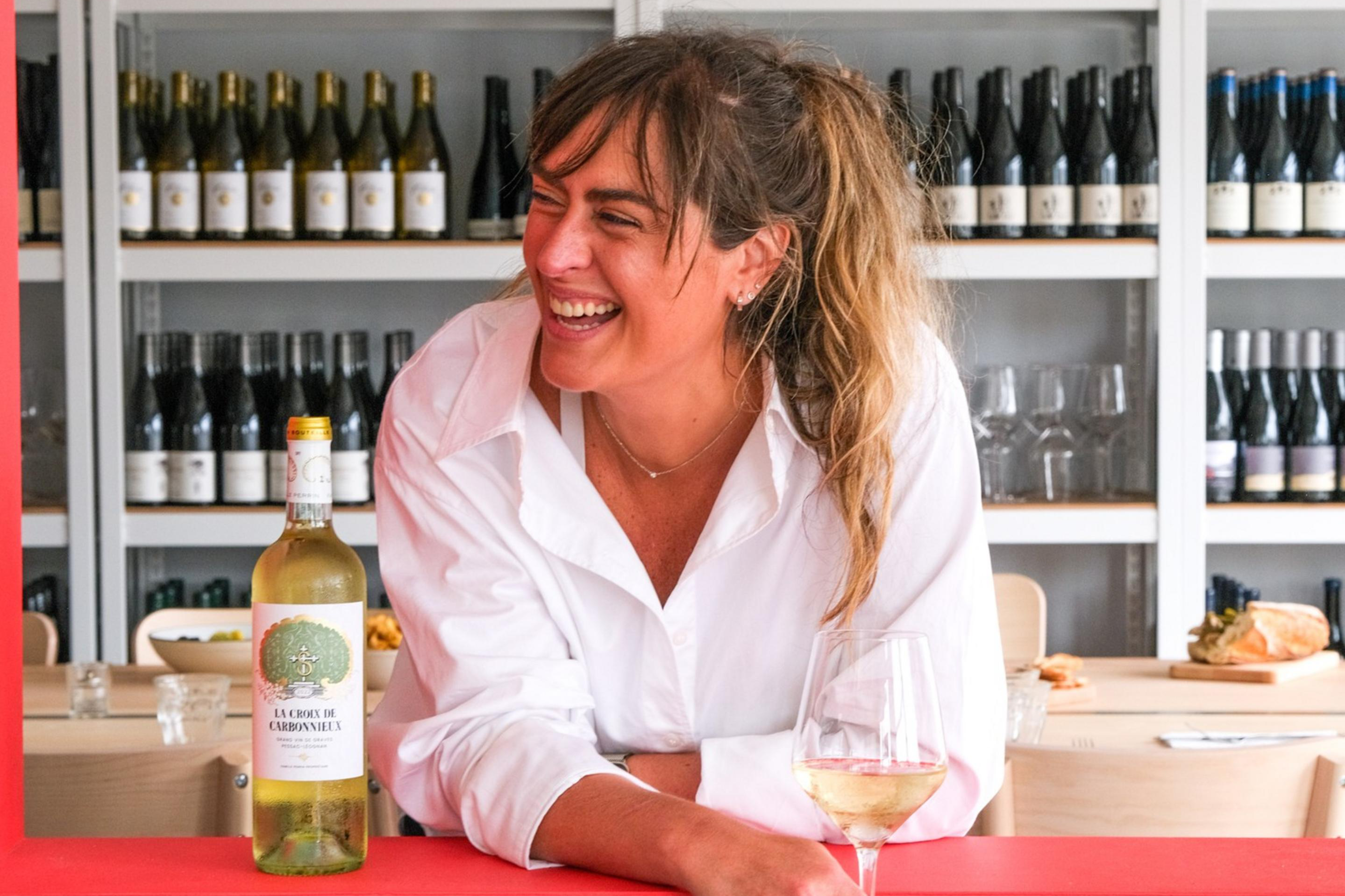 A smiling woman in a white shirt holds a glass of white wine, leaning on a counter with a blue and red backdrop. Shelves of wine bottles and a mermaid painting are behind her.