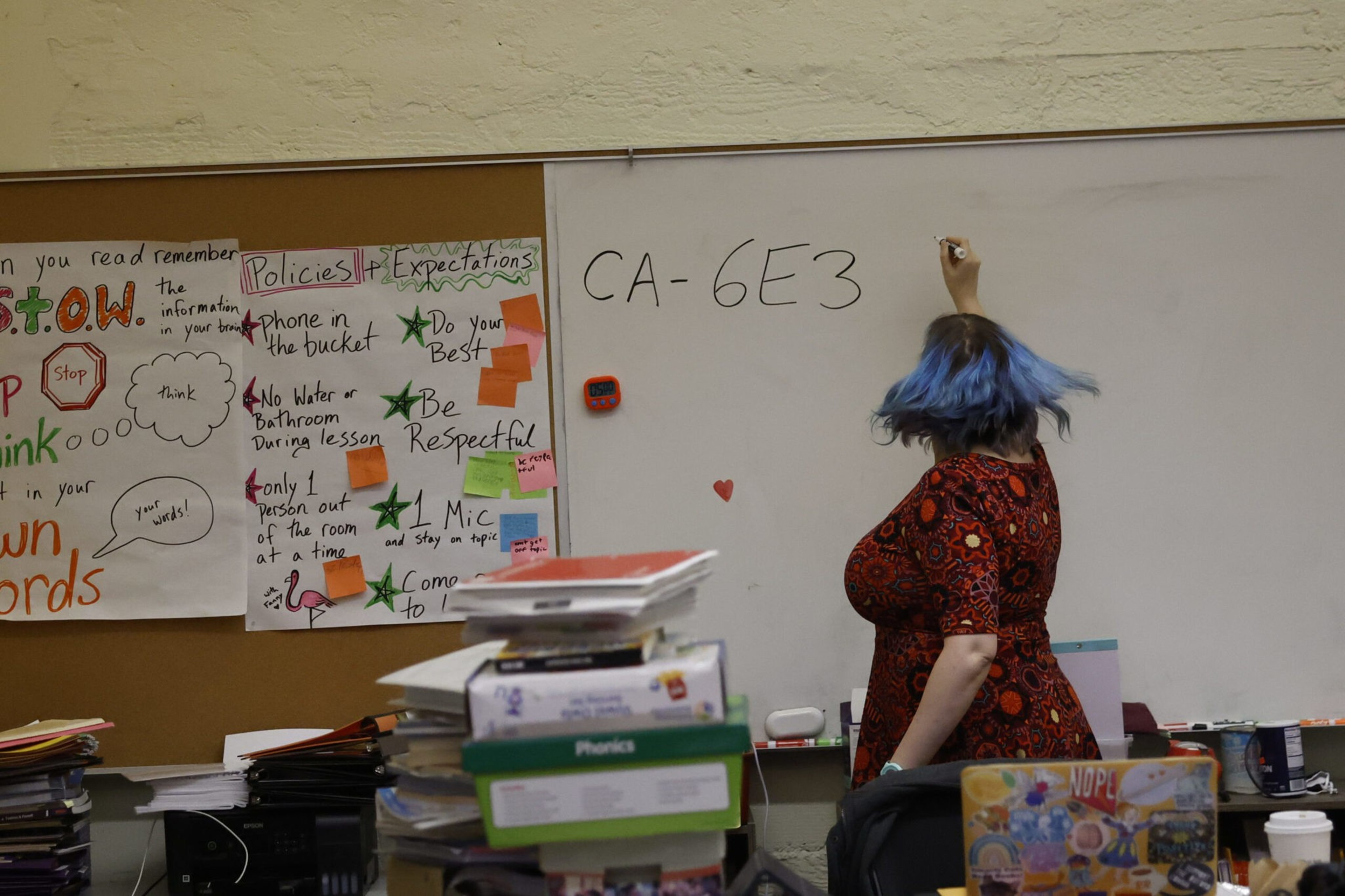 A teacher with blue hair writes "CA-6E3" on a whiteboard in a classroom filled with stacked books and colorful posters on policies and expectations.