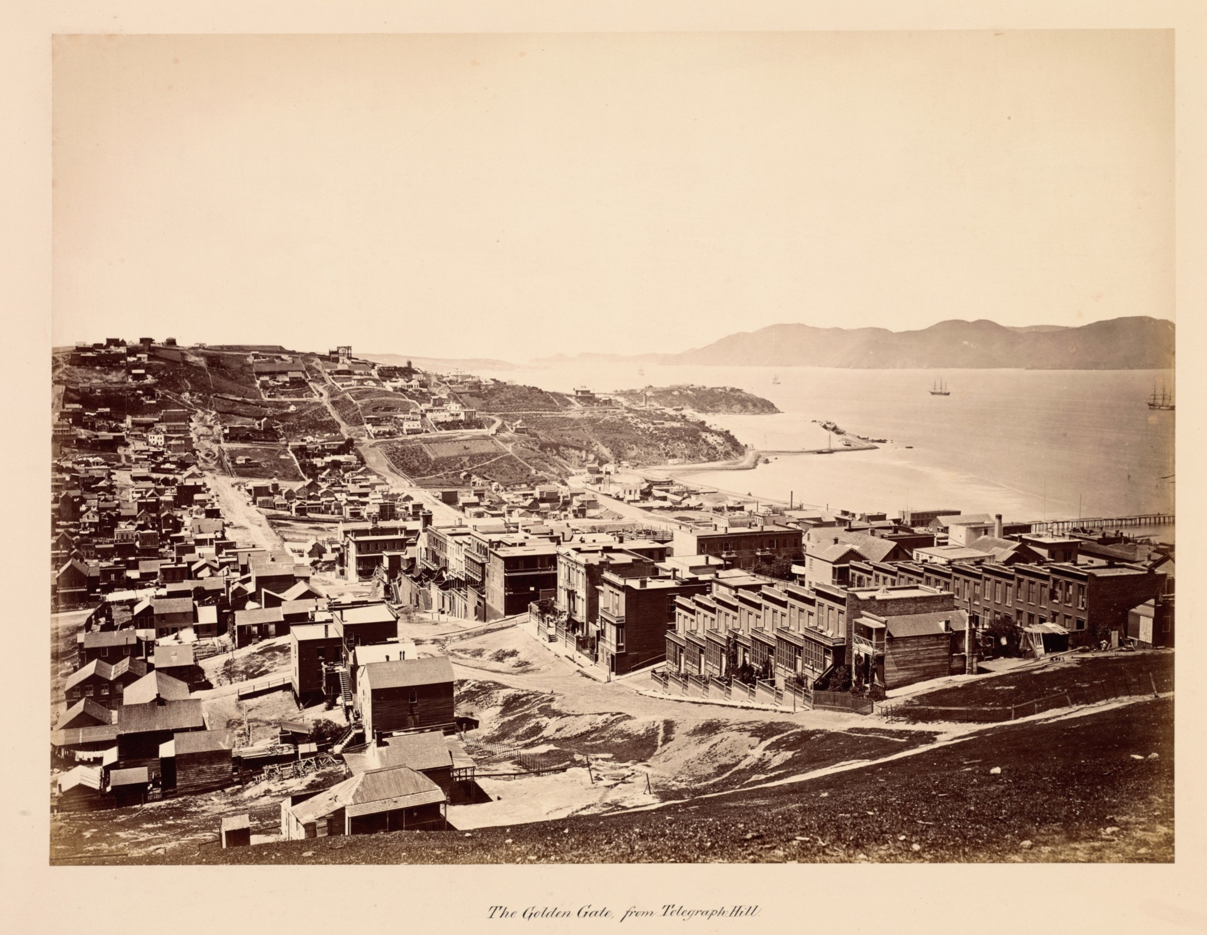 A hilly coastal town with densely packed buildings and dirt roads overlooks a bay with sailing ships and a distant mountain range.