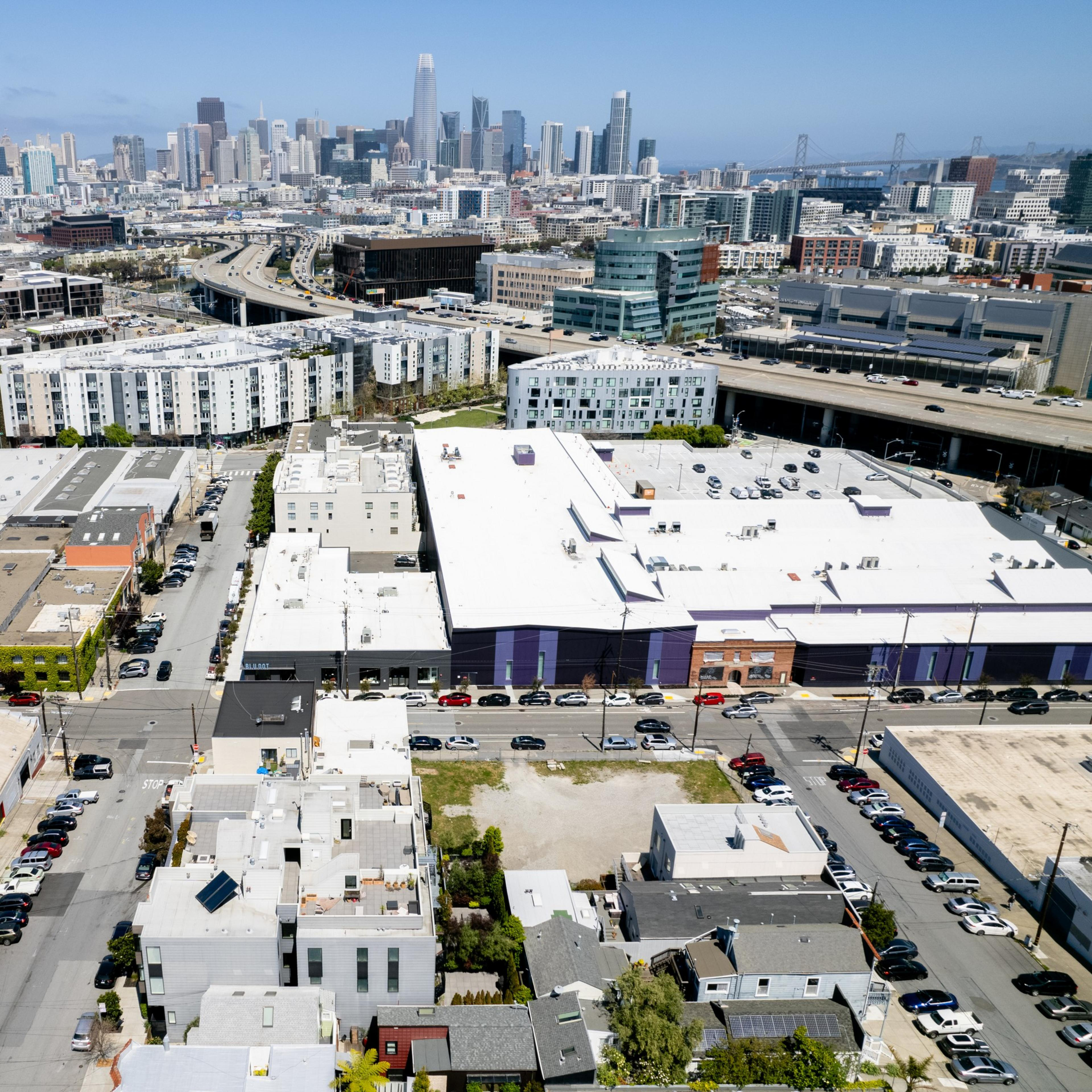 Aerial view of an urban area with buildings, roads, and a distant city skyline under a clear sky.