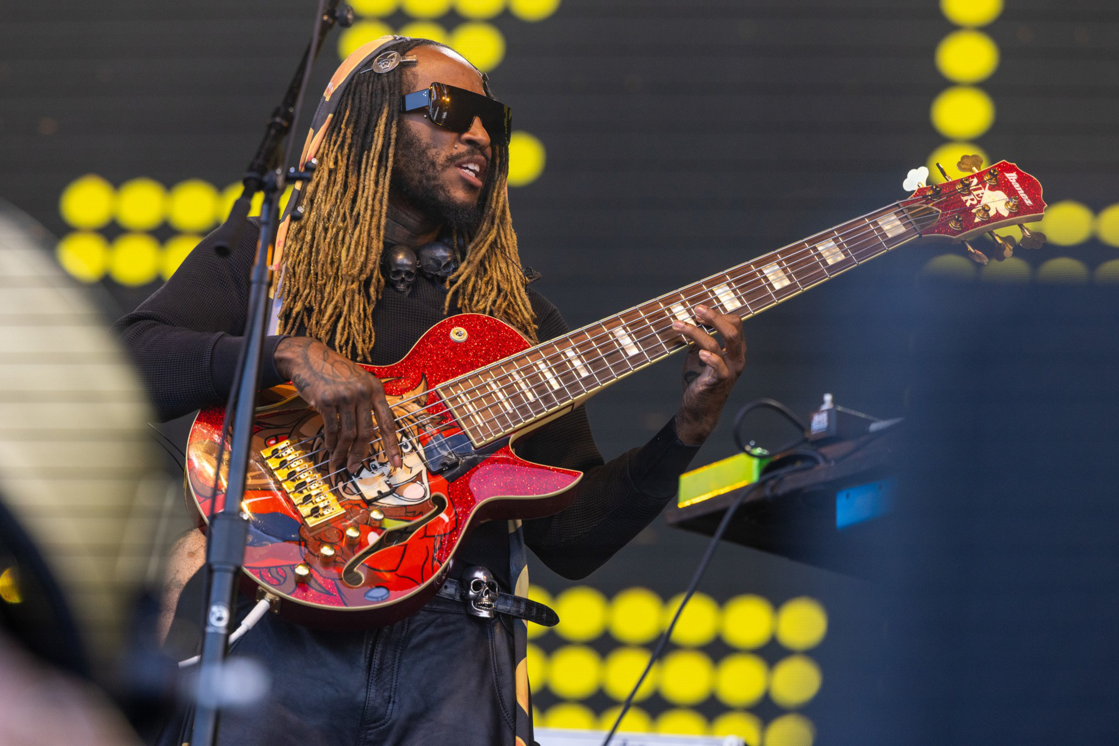 A musician wearing sunglasses and headphones plays a red electric bass guitar with long dreadlocks and a black outfit on stage.