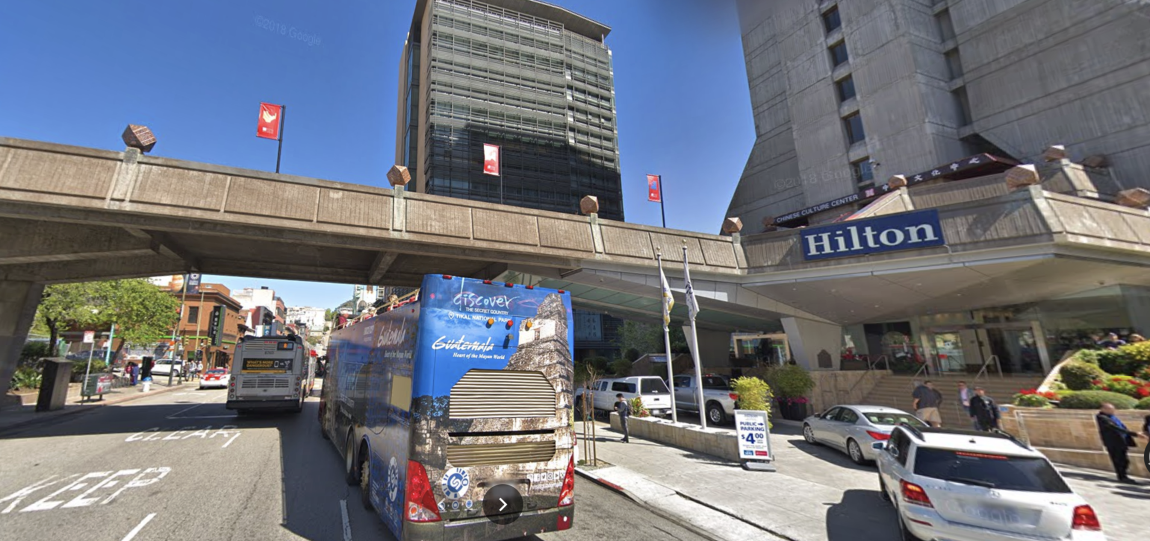 A busy urban street with a Hilton hotel entrance on the right, buses and cars on the road, and a pedestrian overpass overhead under a clear blue sky.