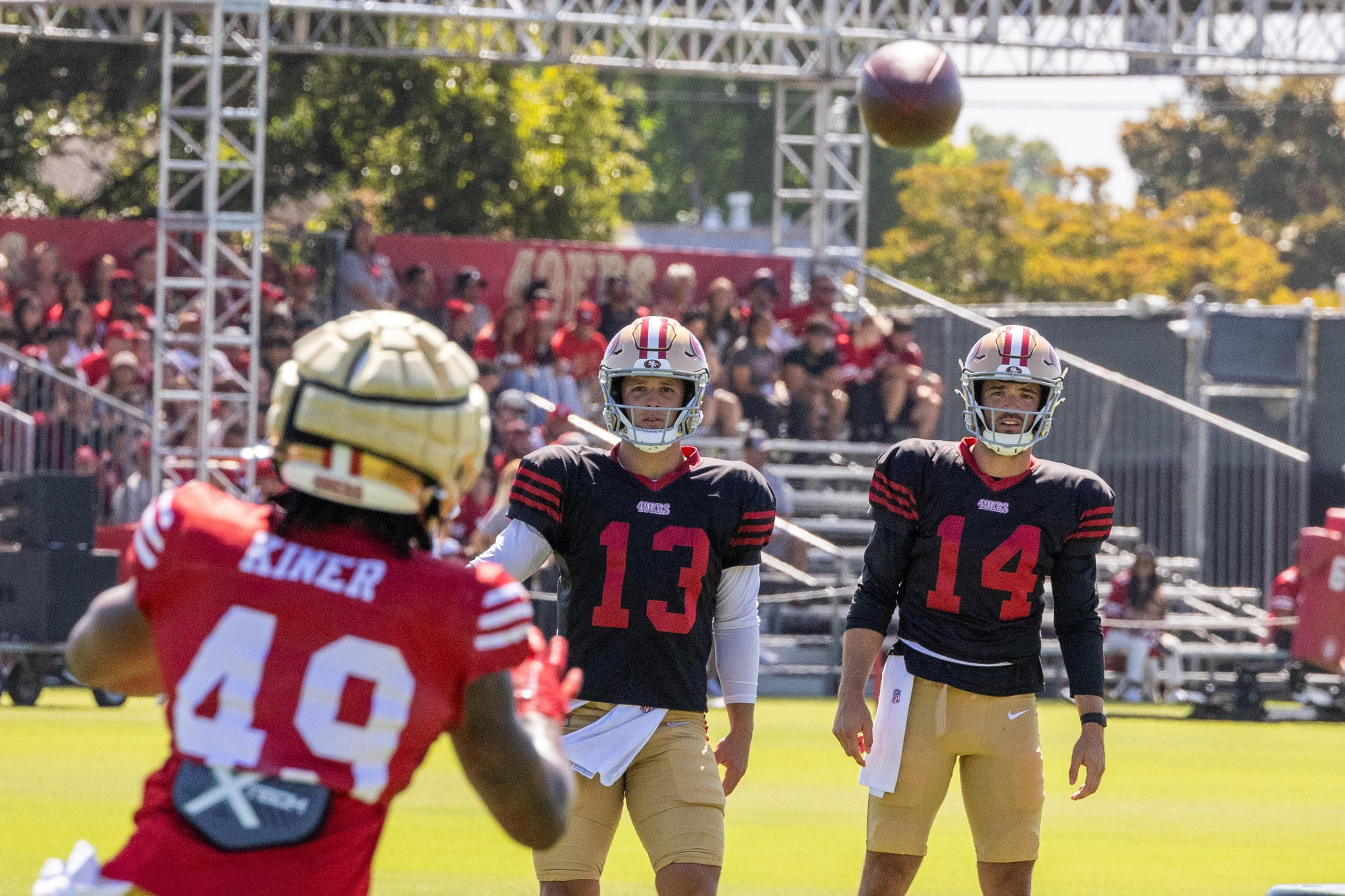 Football players are on a sunny field during practice. Two players in black jerseys with red numbers focus on the ball. Another player in a red jersey is turned away.