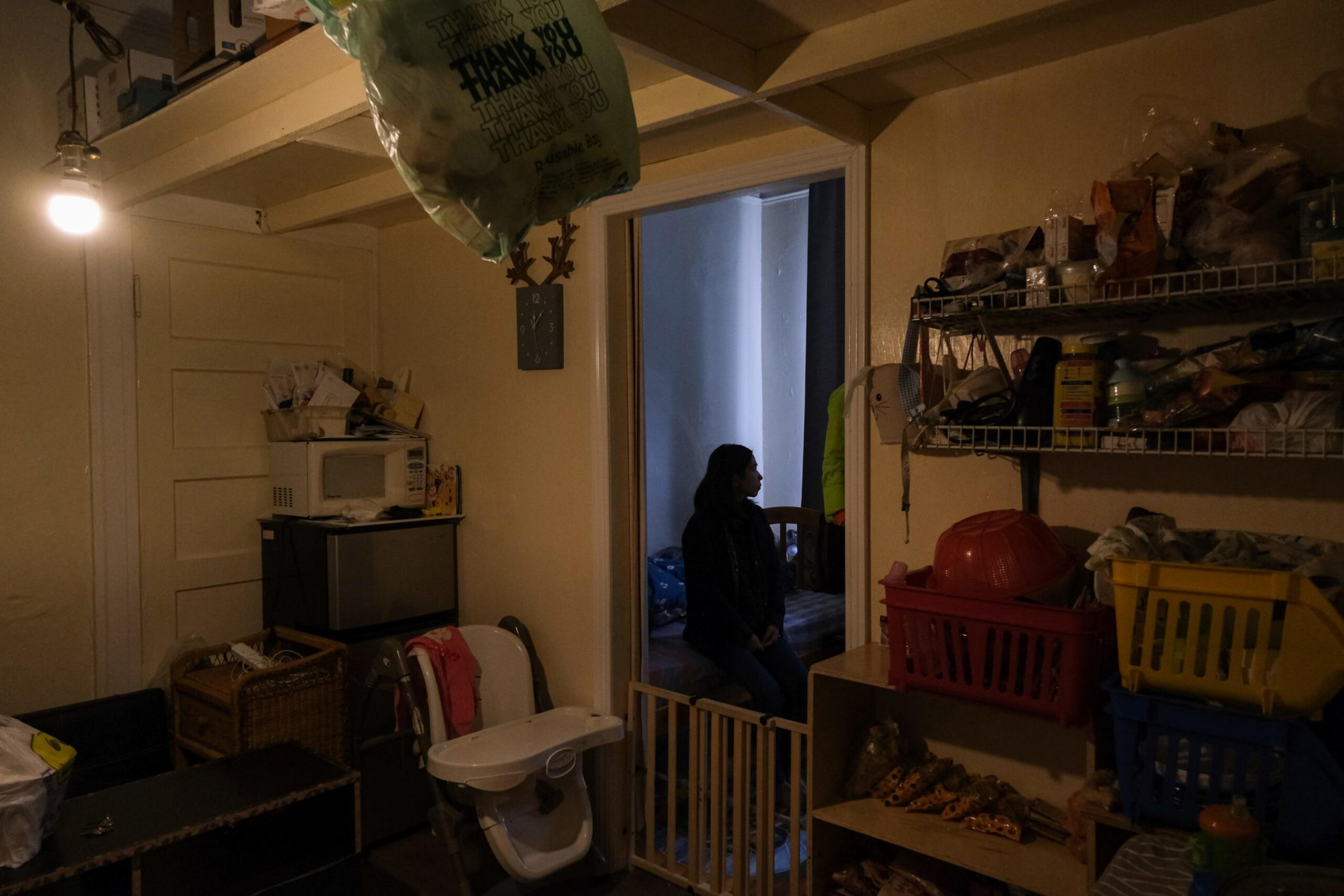A dimly lit room with storage shelves, a high chair, a microwave, and a person sitting in a doorway with a baby gate in front.