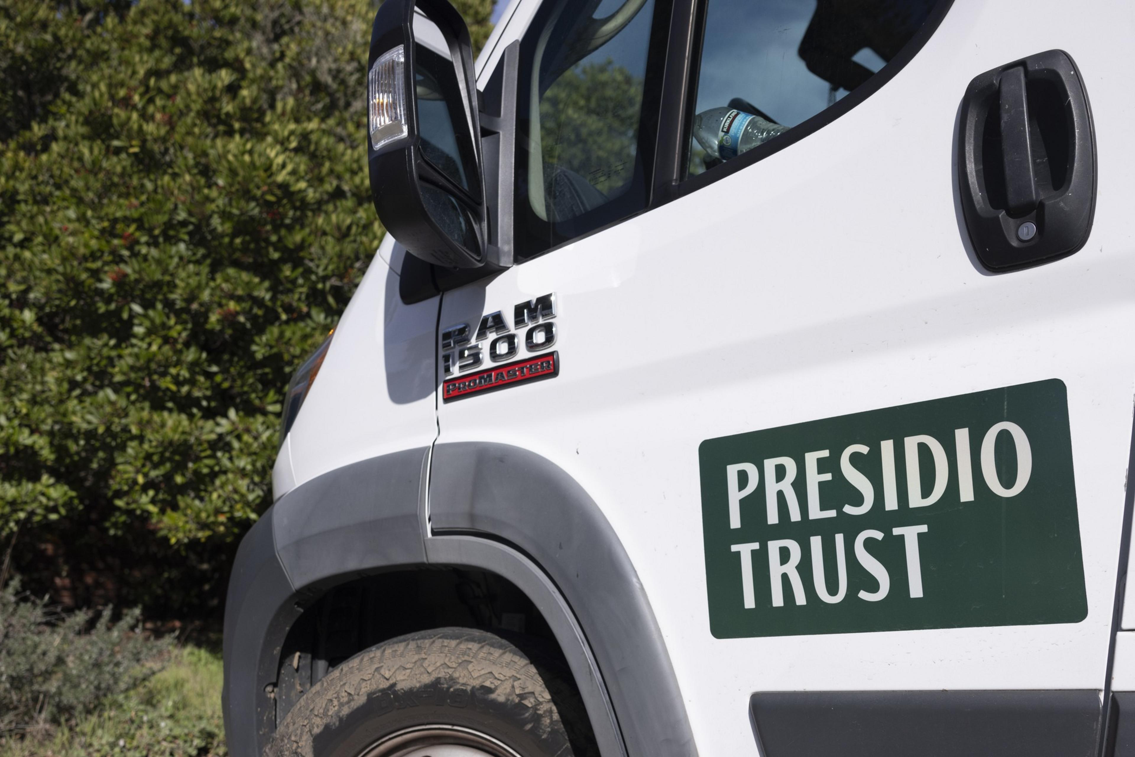 A white RAM 1500 van with a “Presidio Trust” sign on the door is parked near green bushes in daylight.