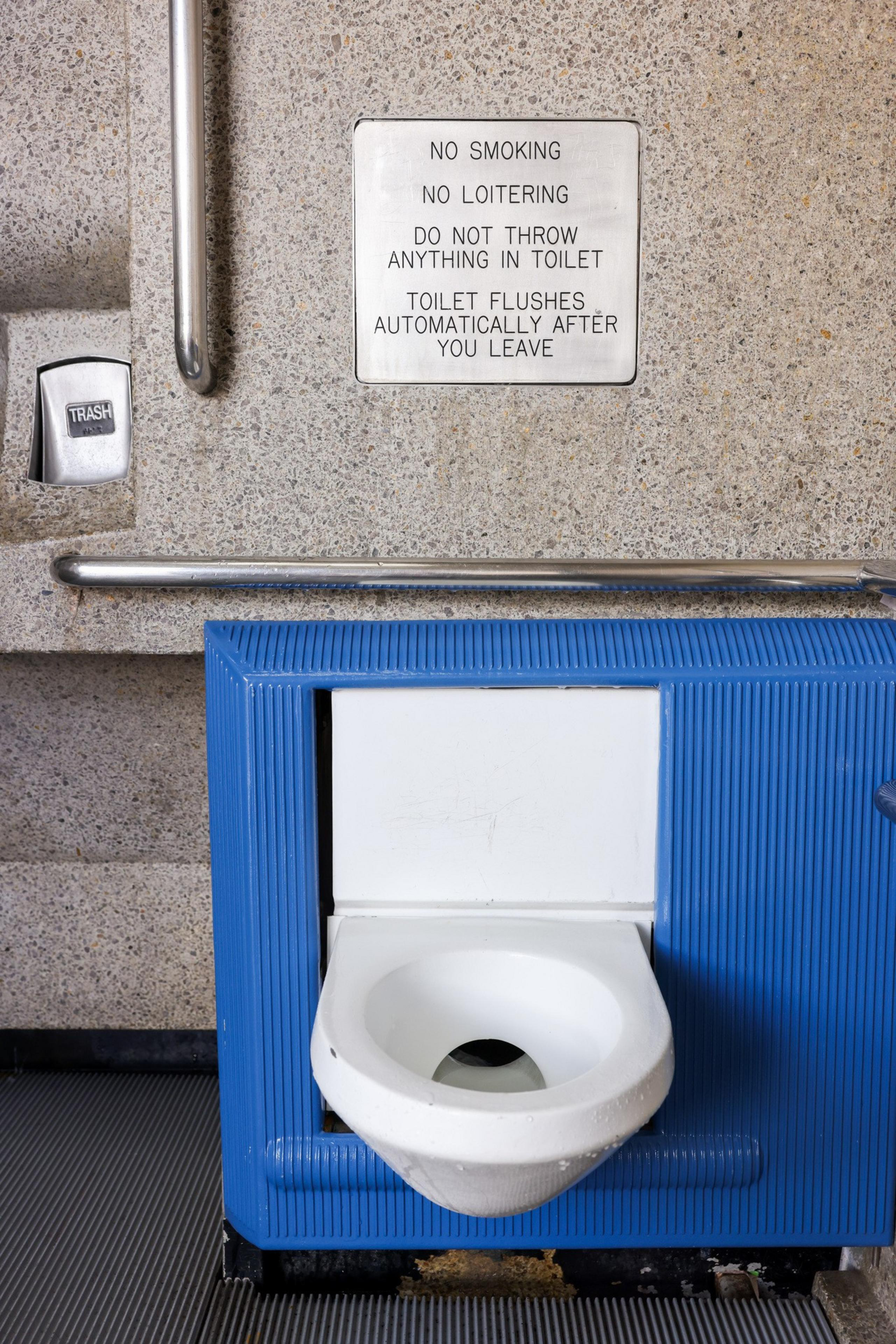 A restroom wall with a sign warning against smoking and loitering. A blue-framed toilet, a trash bin, and metal handrails are visible. The toilet flushes automatically.