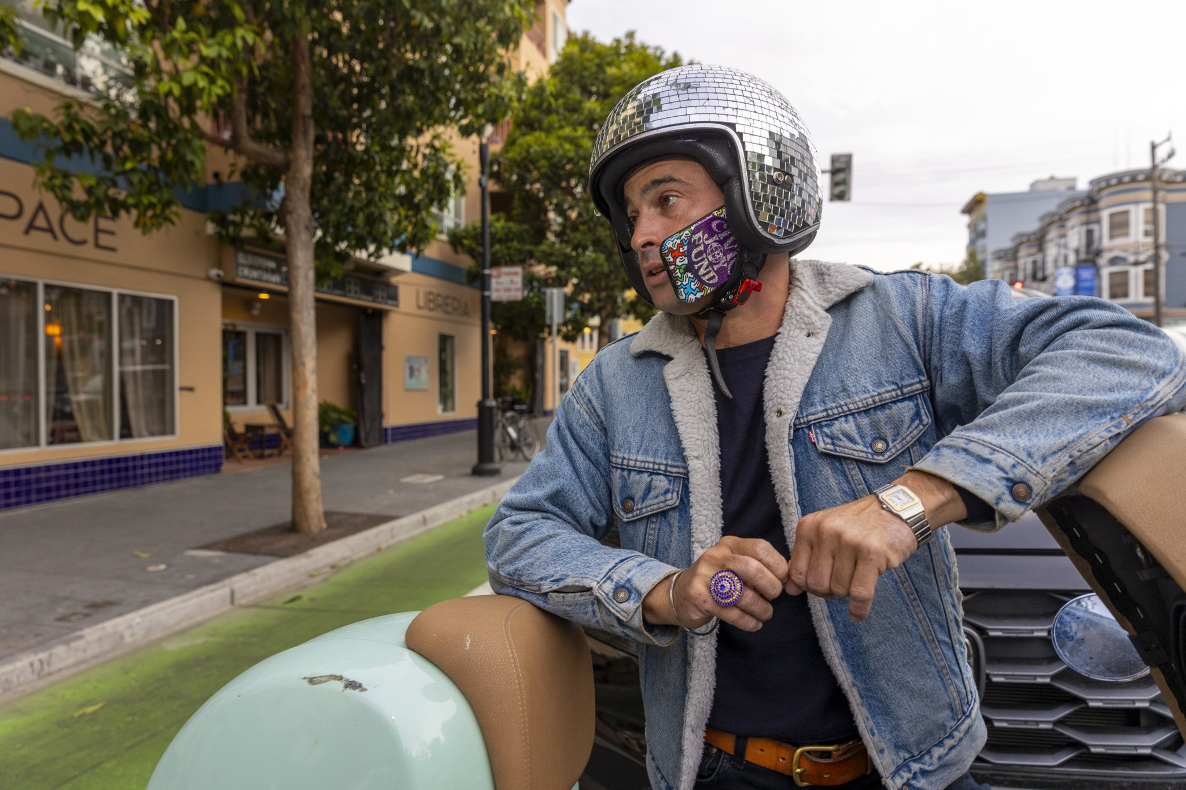 A man wearing a disco ball-style helmet and denim jacket leans on a pastel scooter seat on a city street with trees and buildings nearby.