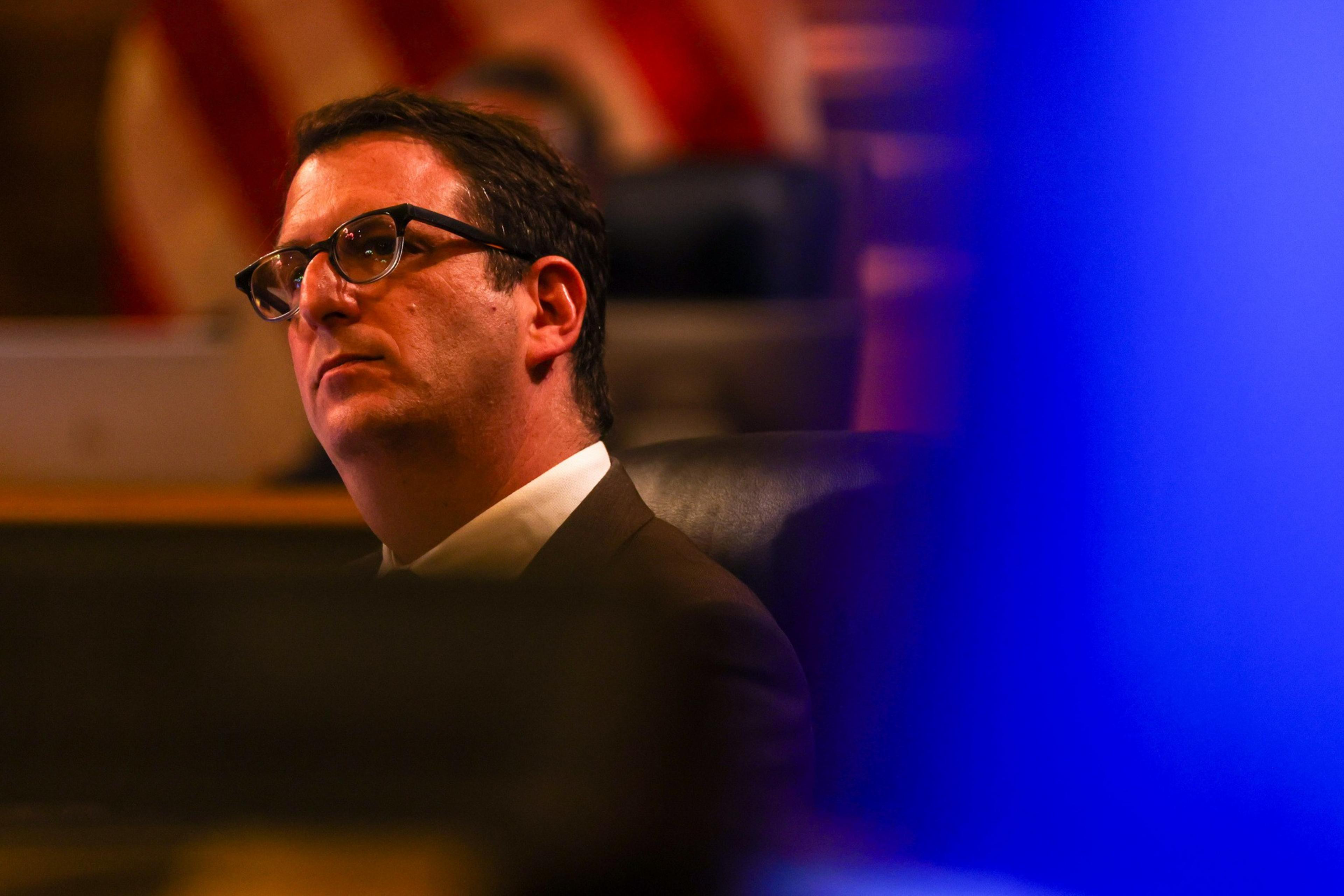A man with glasses is seated, gazing attentively to the side in a dimly lit setting. In the background, there's a blurred American flag and a bright blue area.