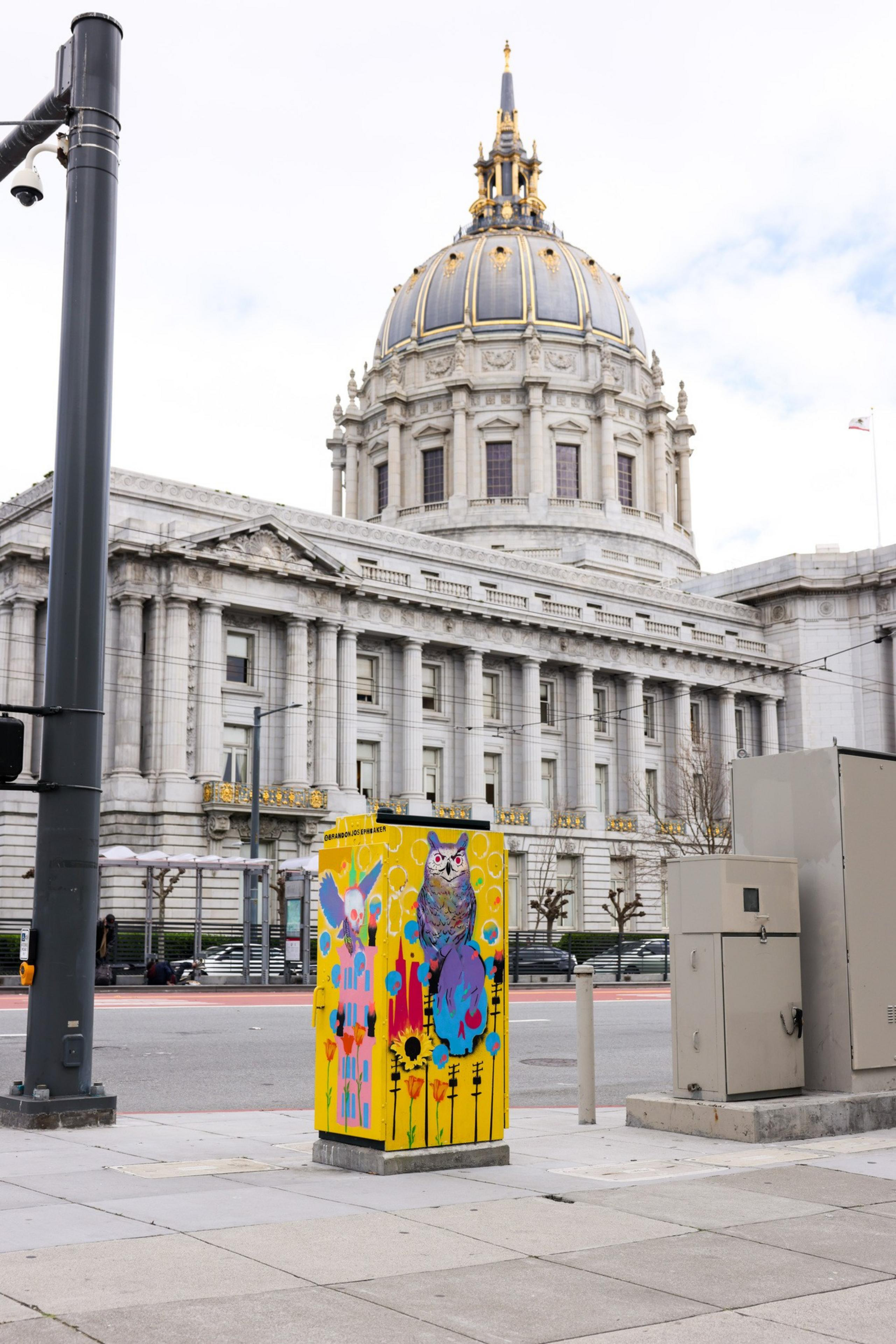 A decorated utility box with colorful owls and flowers is in front of an ornate building with a large dome and intricate architecture.