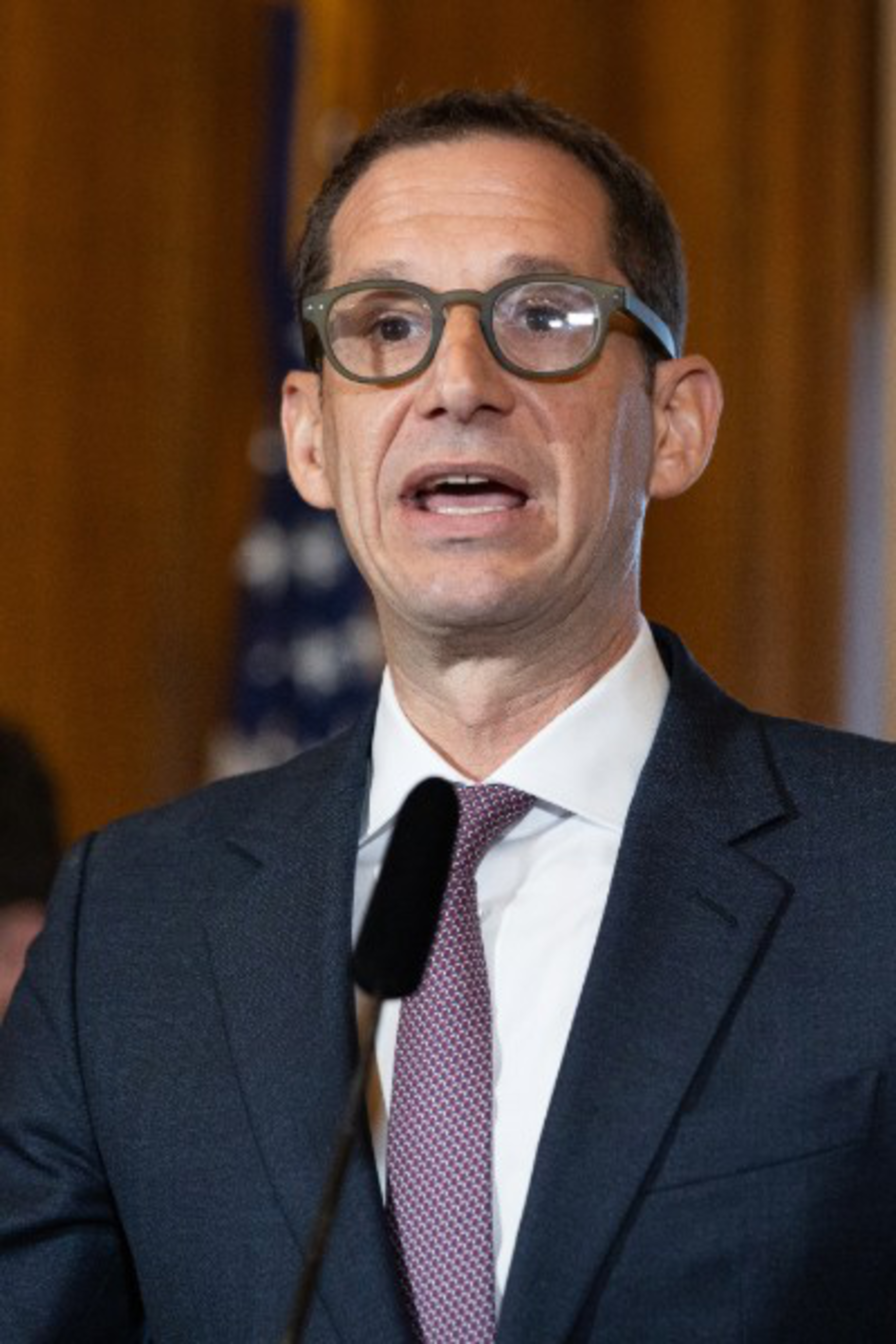 In a composite photo, Mayor Daniel Lurie wears glasses and speaks at a podium on the left, while Donald Trump looks out from behind a podium on the right.