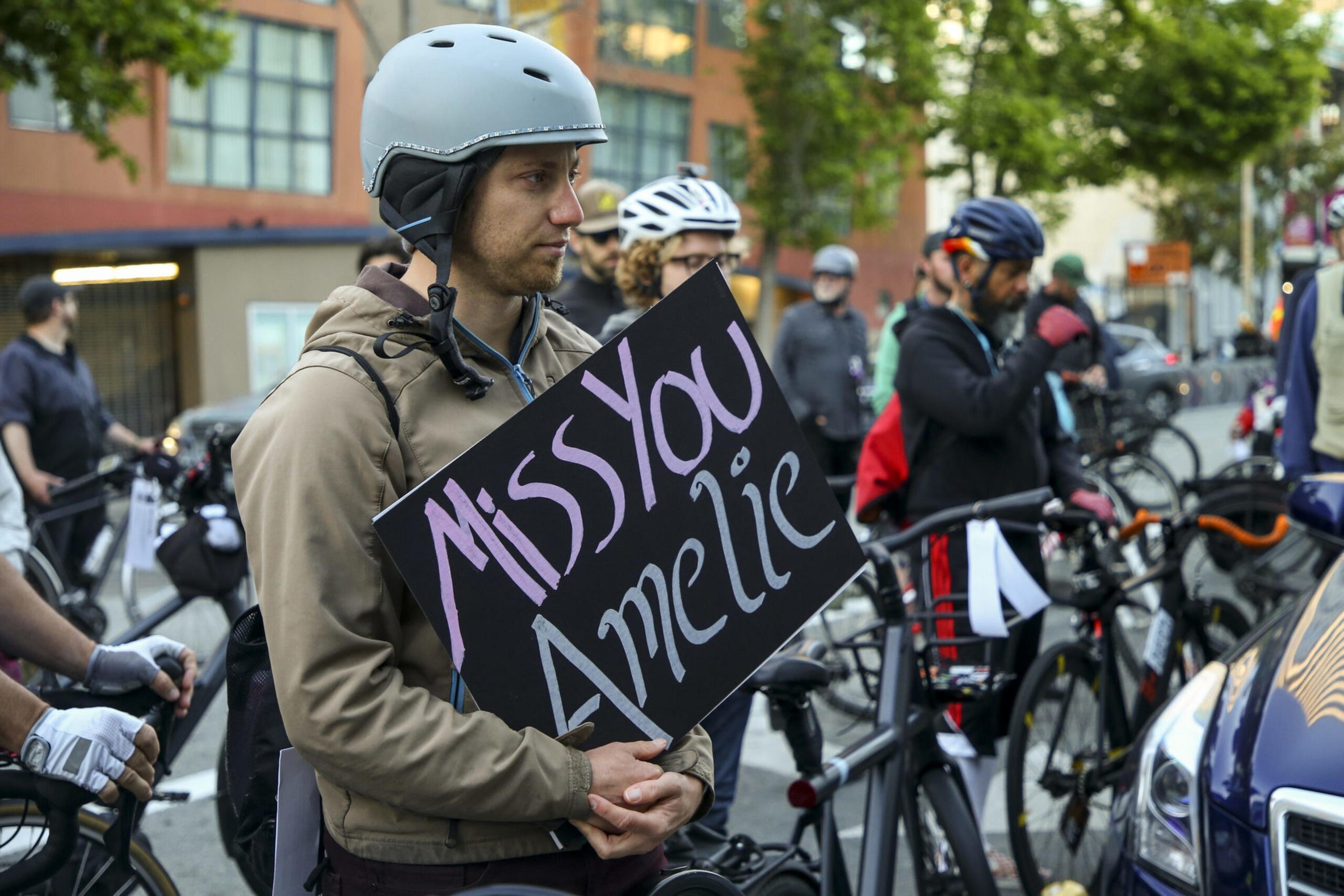 A person wearing a helmet and jacket holds a sign reading "Miss You Amelie" among a group of cyclists on a city street.