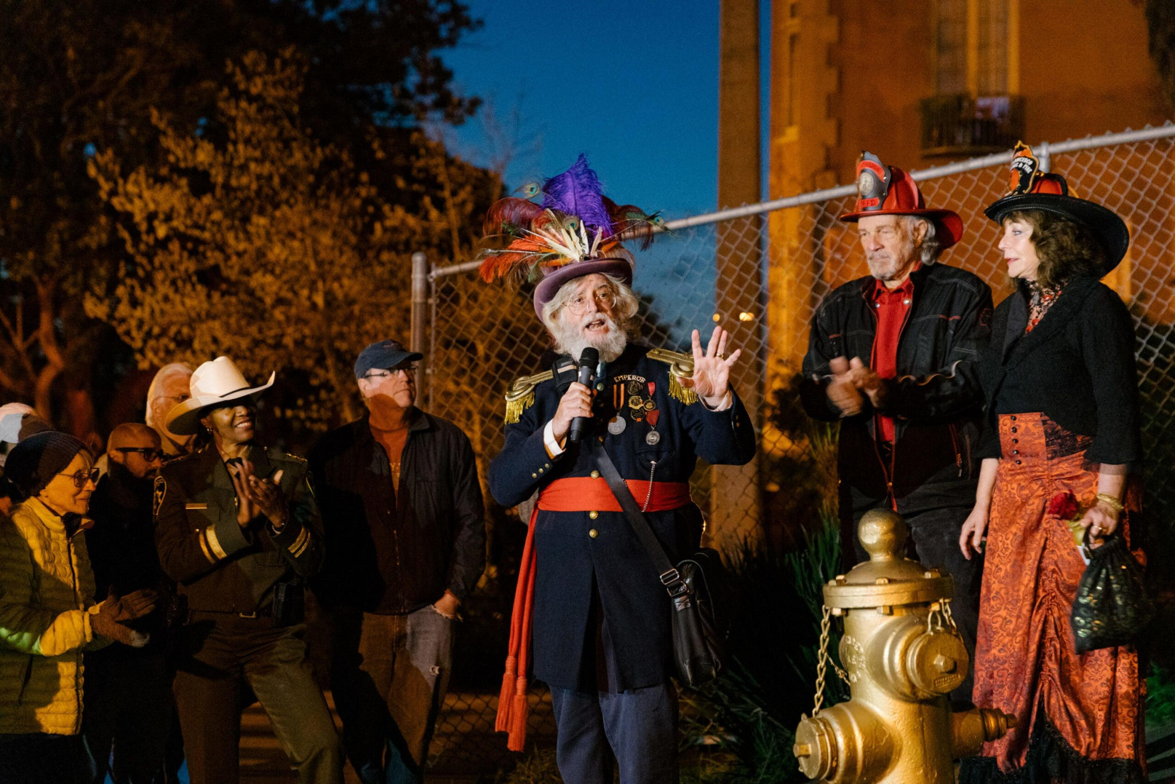 An elderly man in a colorful feathered hat and military-style coat speaks into a microphone as a small crowd, some in costumes, listens and claps.