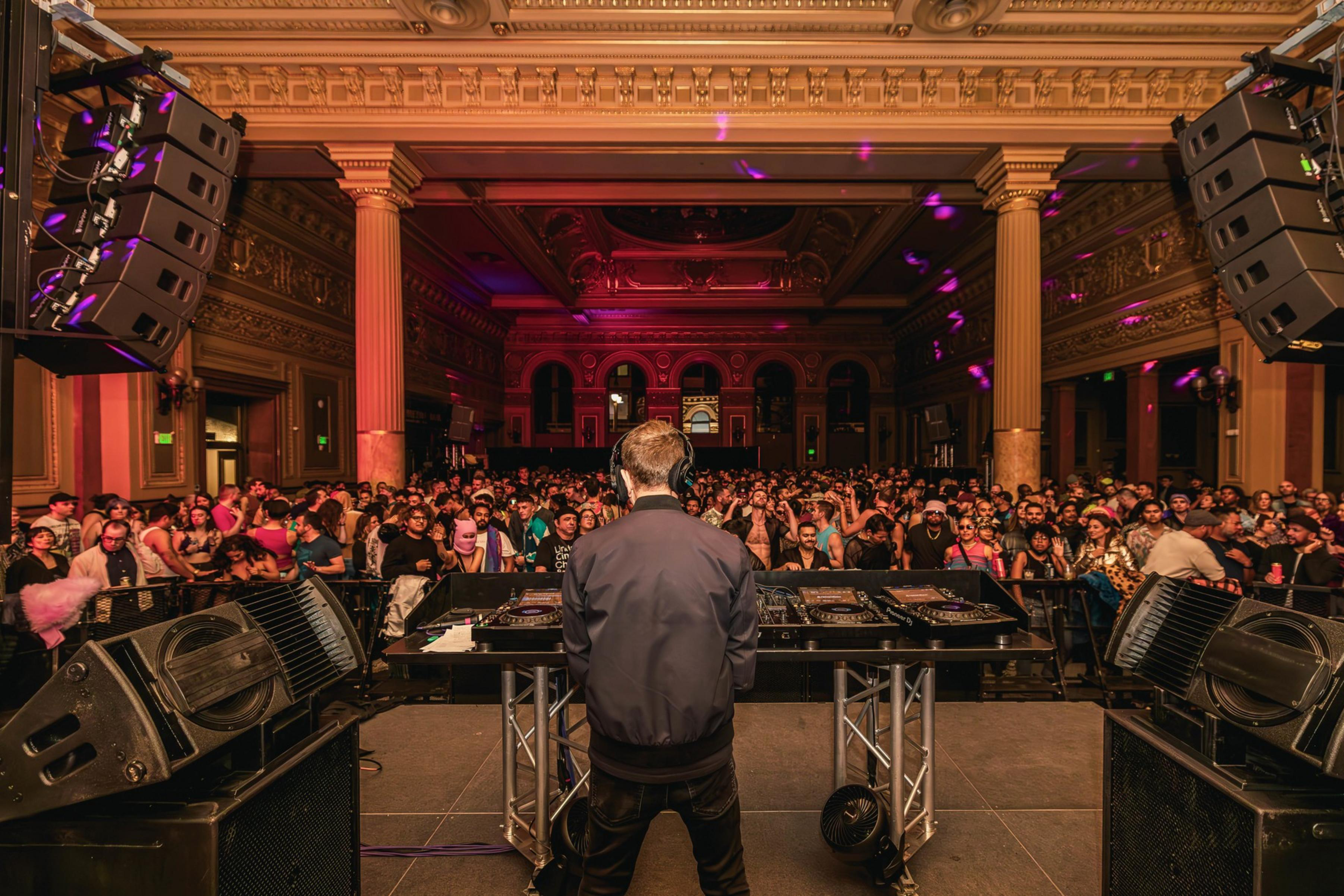 A DJ, seen from behind, performs on stage with large speakers in an opulent hall filled with a vibrant crowd, under colorful lighting, inside a grand, ornate building.