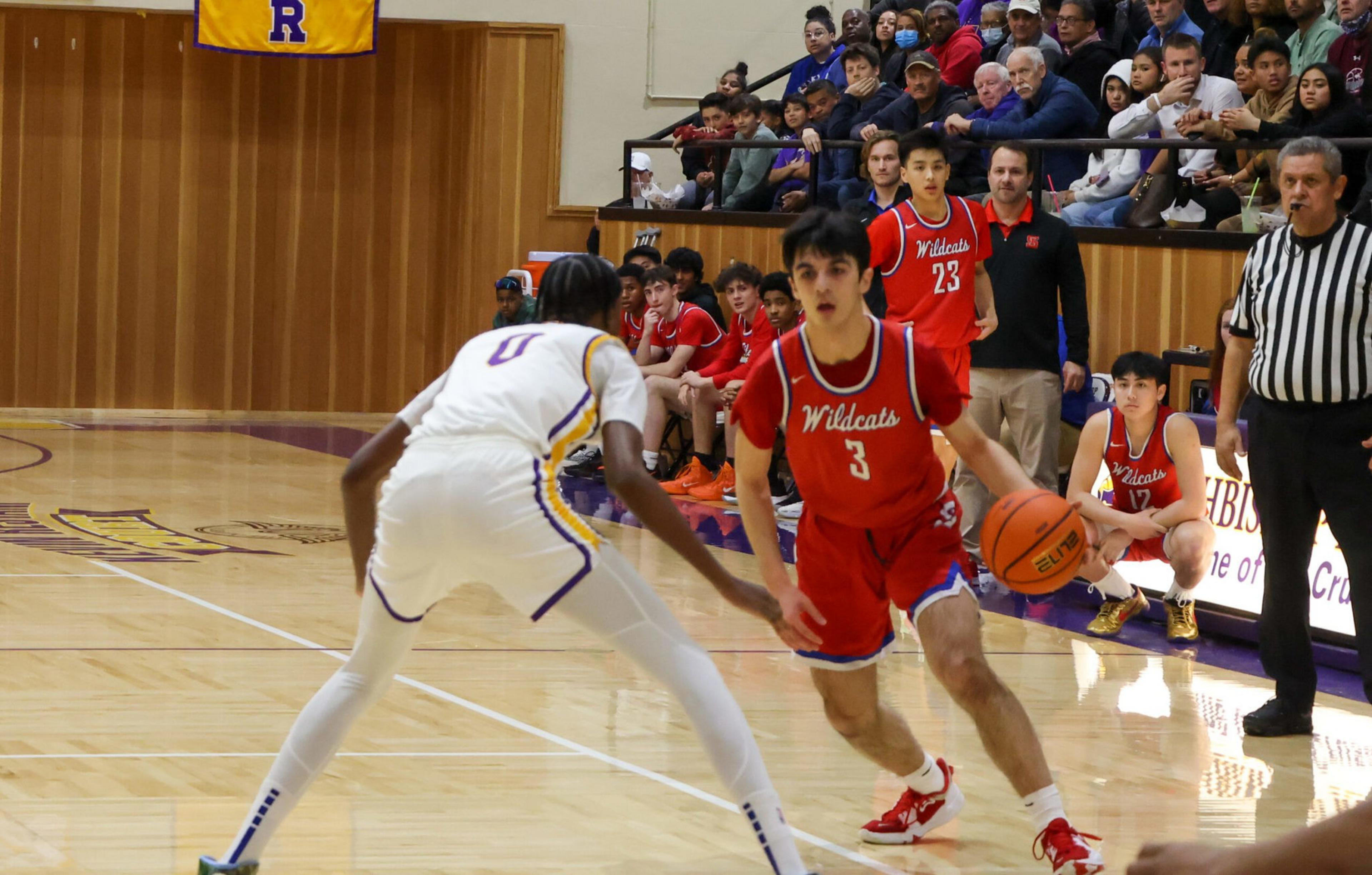 A basketball player in a red Wildcats uniform dribbles while being defended by a player in a white uniform, with teammates, coach, and audience watching.