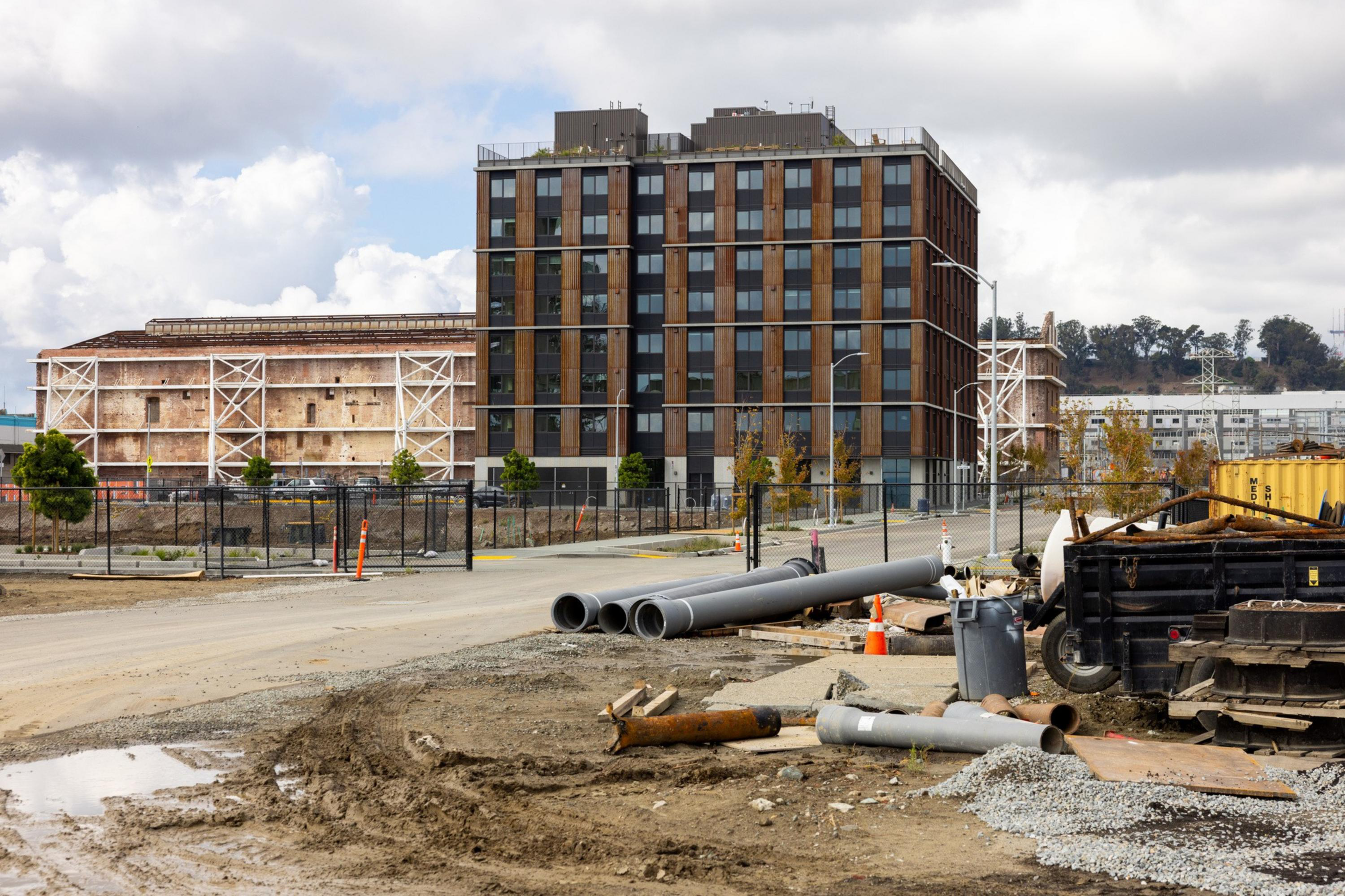 A construction site with dirt, gravel, pipes, and equipment sits in front of a modern multi-story building and an older warehouse.