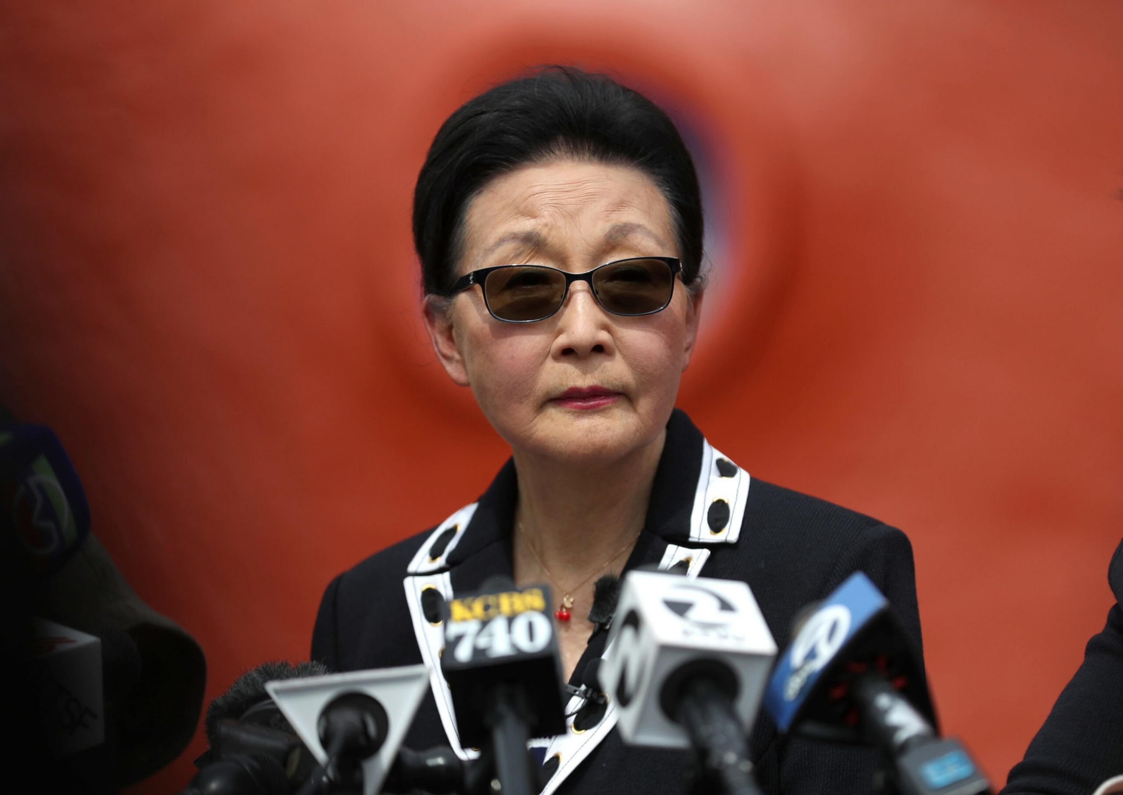 A woman wearing dark sunglasses and a black-and-white jacket speaks at a press conference with multiple microphones in front of her.