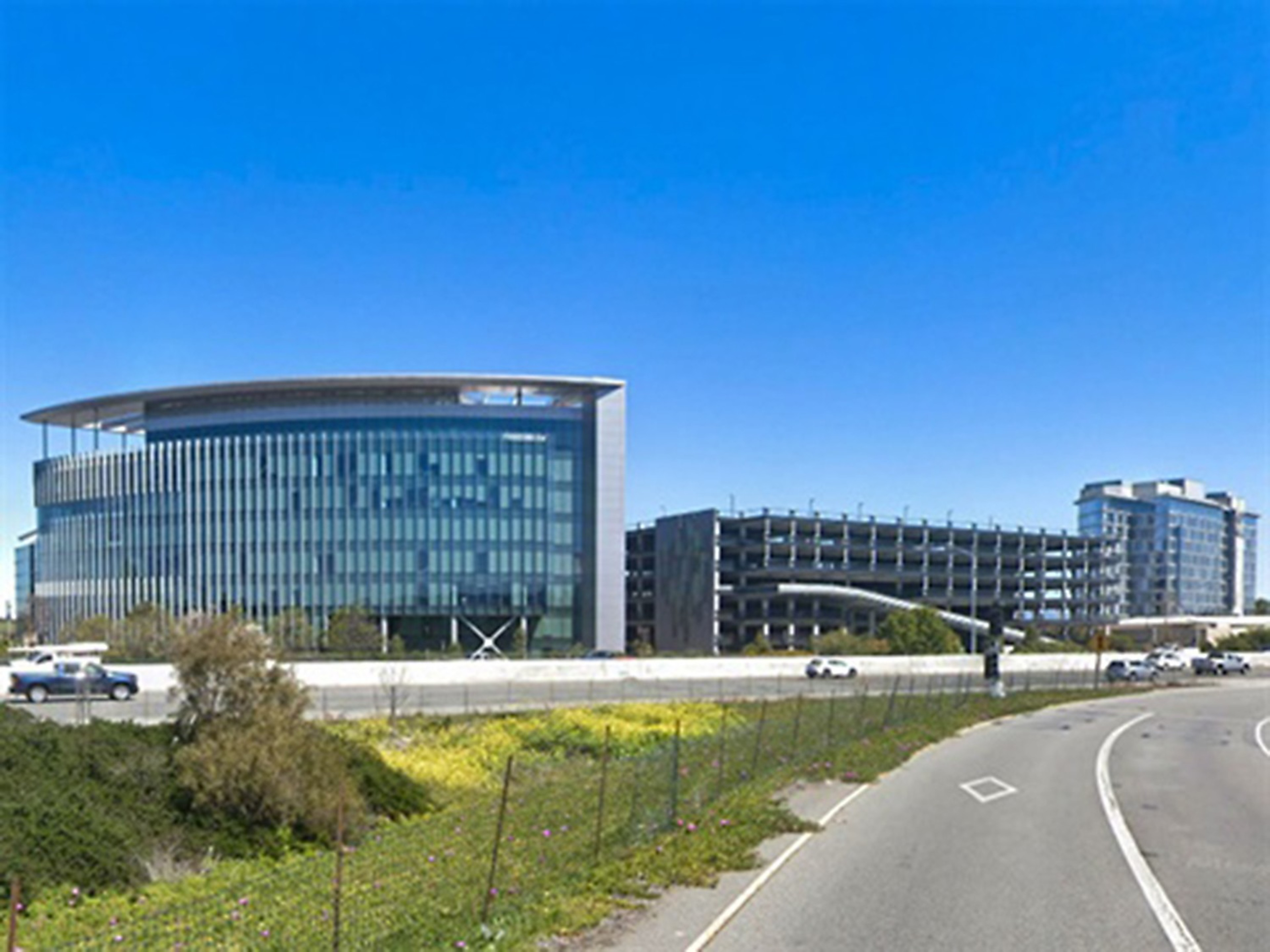 The image shows a modern glass building with vertical lines, a multi-story parking structure, and another glass building, set against a clear blue sky. A road curves in the foreground.