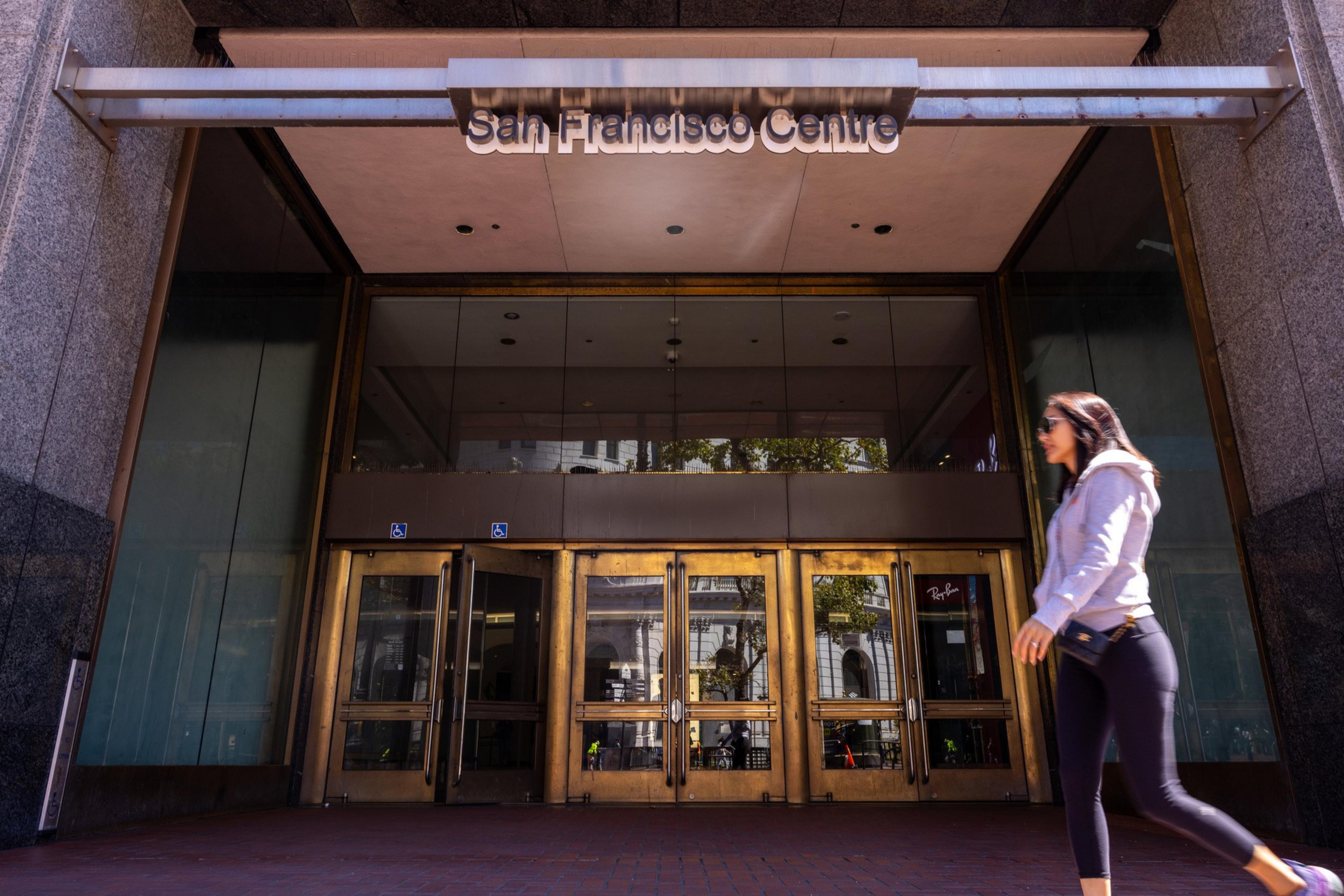 A woman in sunglasses, a light hoodie, and black leggings walks past the glass and bronze entrance doors of the San Francisco Centre.