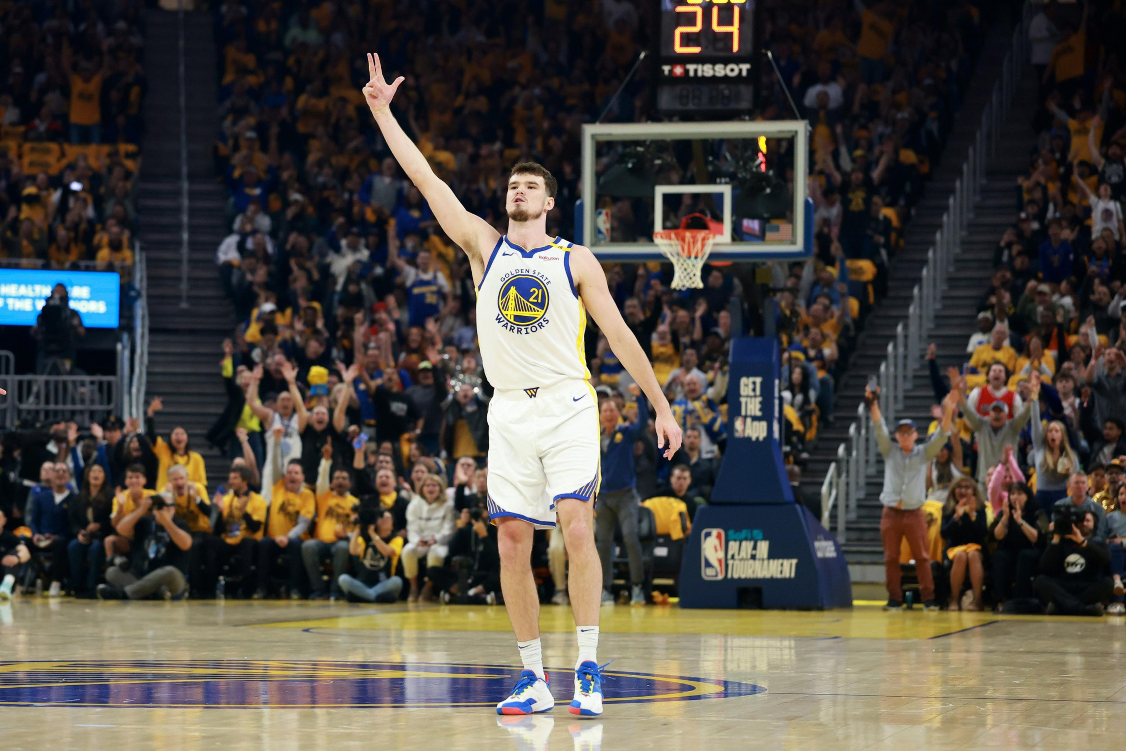 A basketball player in a Golden State Warriors uniform holds up two fingers on a court. The crowd behind him is cheering enthusiastically.