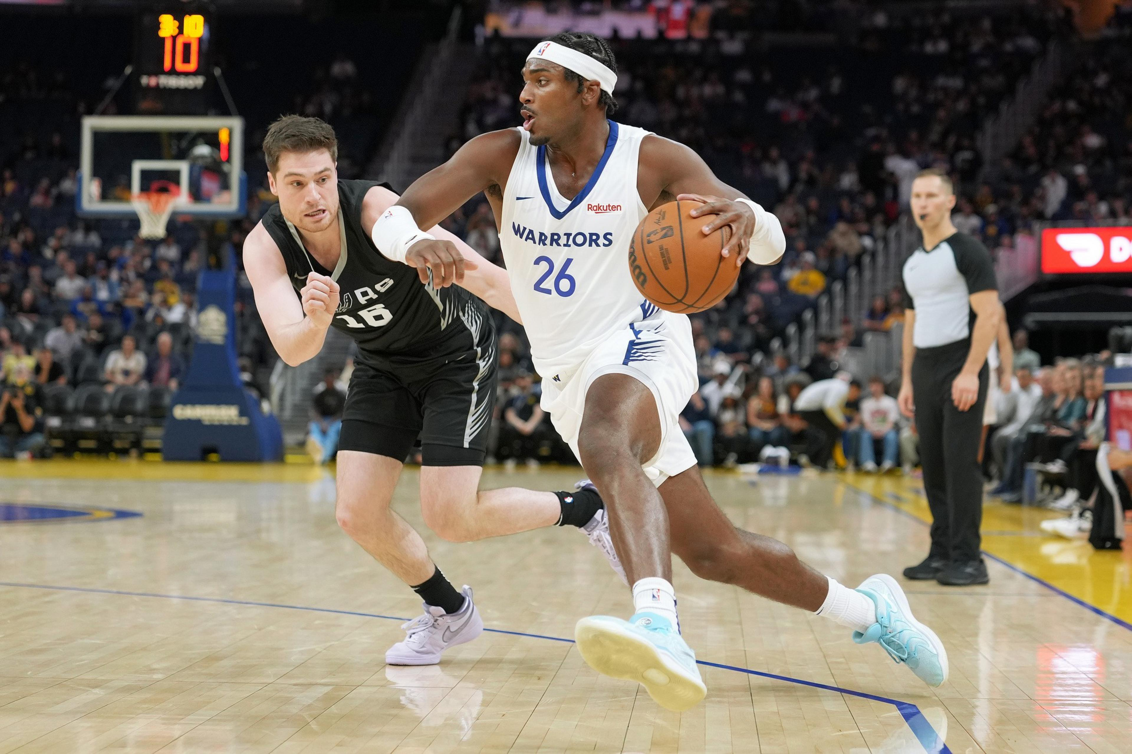 A basketball player in a white &quot;Warriors&quot; uniform is dribbling the ball past a defender in a black uniform. An official and audience are visible in the background.