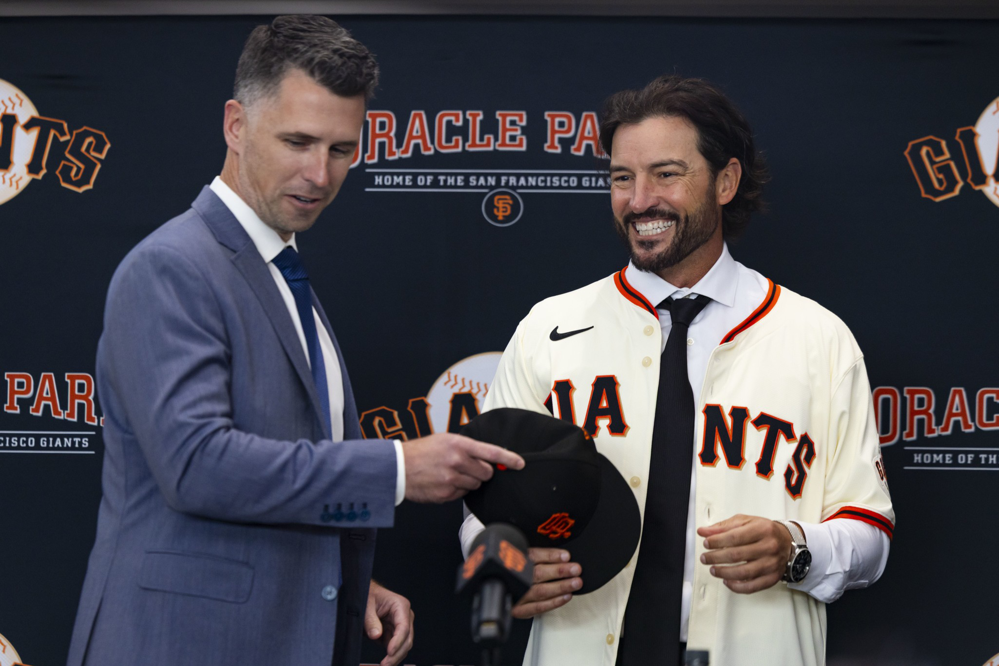 A man in a suit hands a black San Francisco Giants cap to a smiling man wearing a Giants jersey and tie, with an "Oracle Park" backdrop behind them.