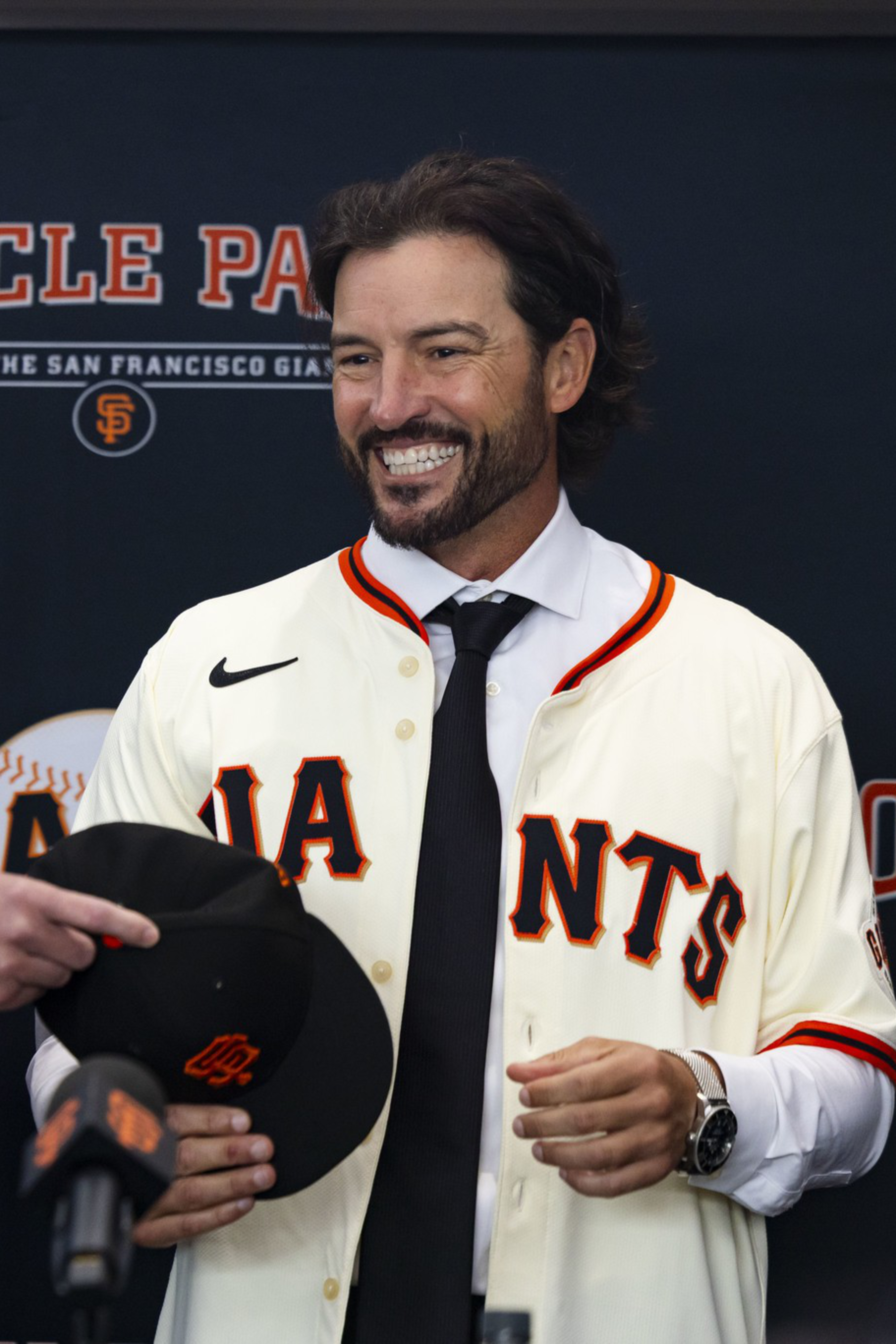 A man in a suit hands a black San Francisco Giants cap to a smiling man wearing a Giants jersey and tie, with an "Oracle Park" backdrop behind them.