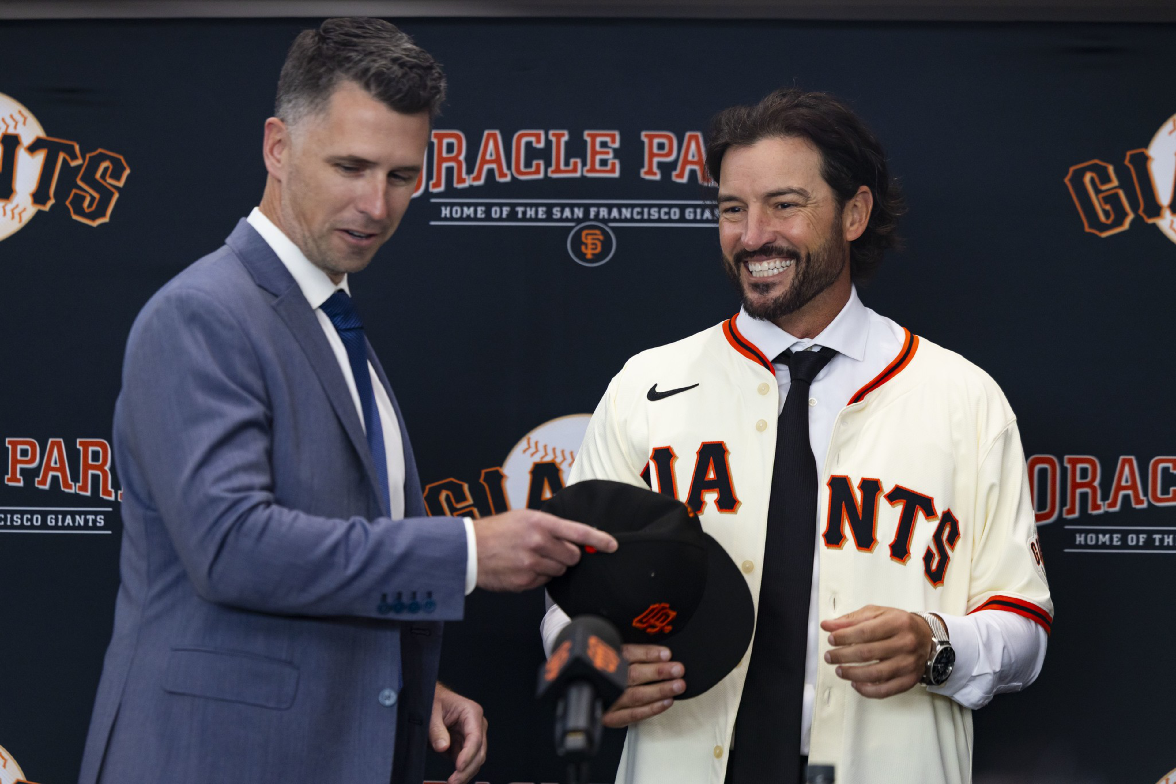 A man in a suit hands a black San Francisco Giants cap to a smiling man wearing a Giants jersey and tie, with an "Oracle Park" backdrop behind them.