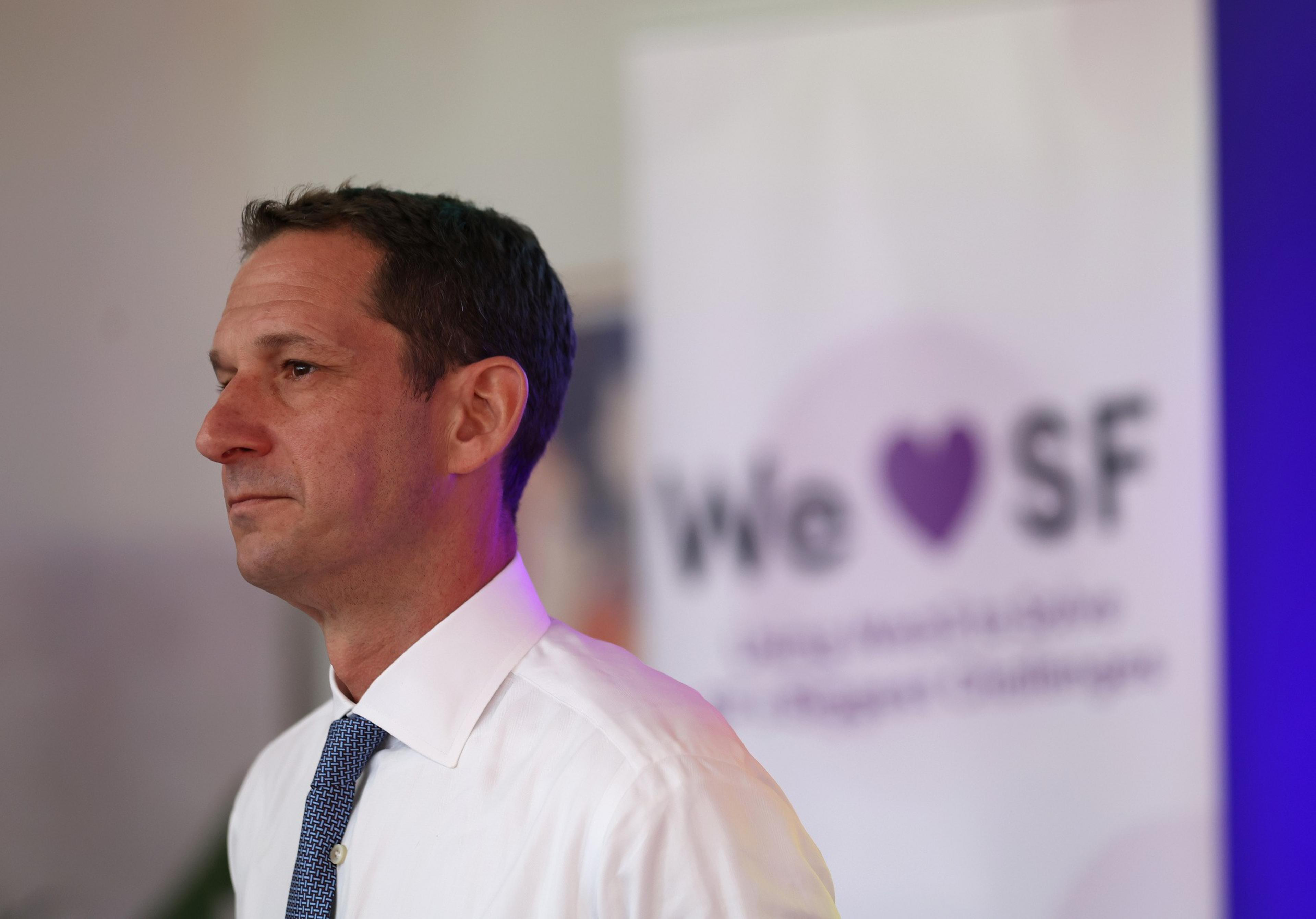 A man in a white shirt and tie stands in profile against a blurred backdrop featuring a banner that says &quot;We ♥ SF&quot; in purple.