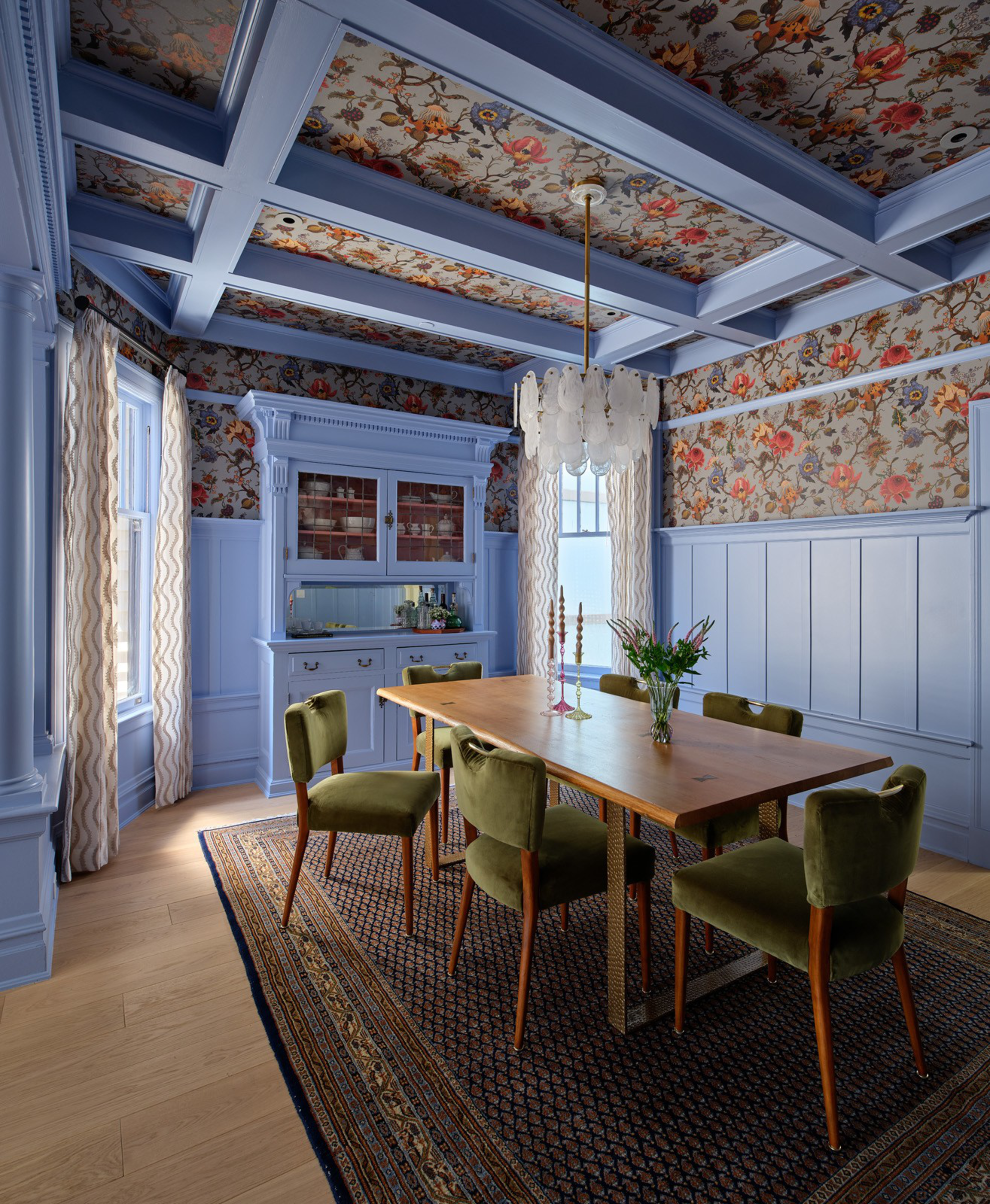 A dining room with lavender wainscoting and floral wallpaper on walls and ceiling, featuring a wooden table, green velvet chairs, and a patterned rug.