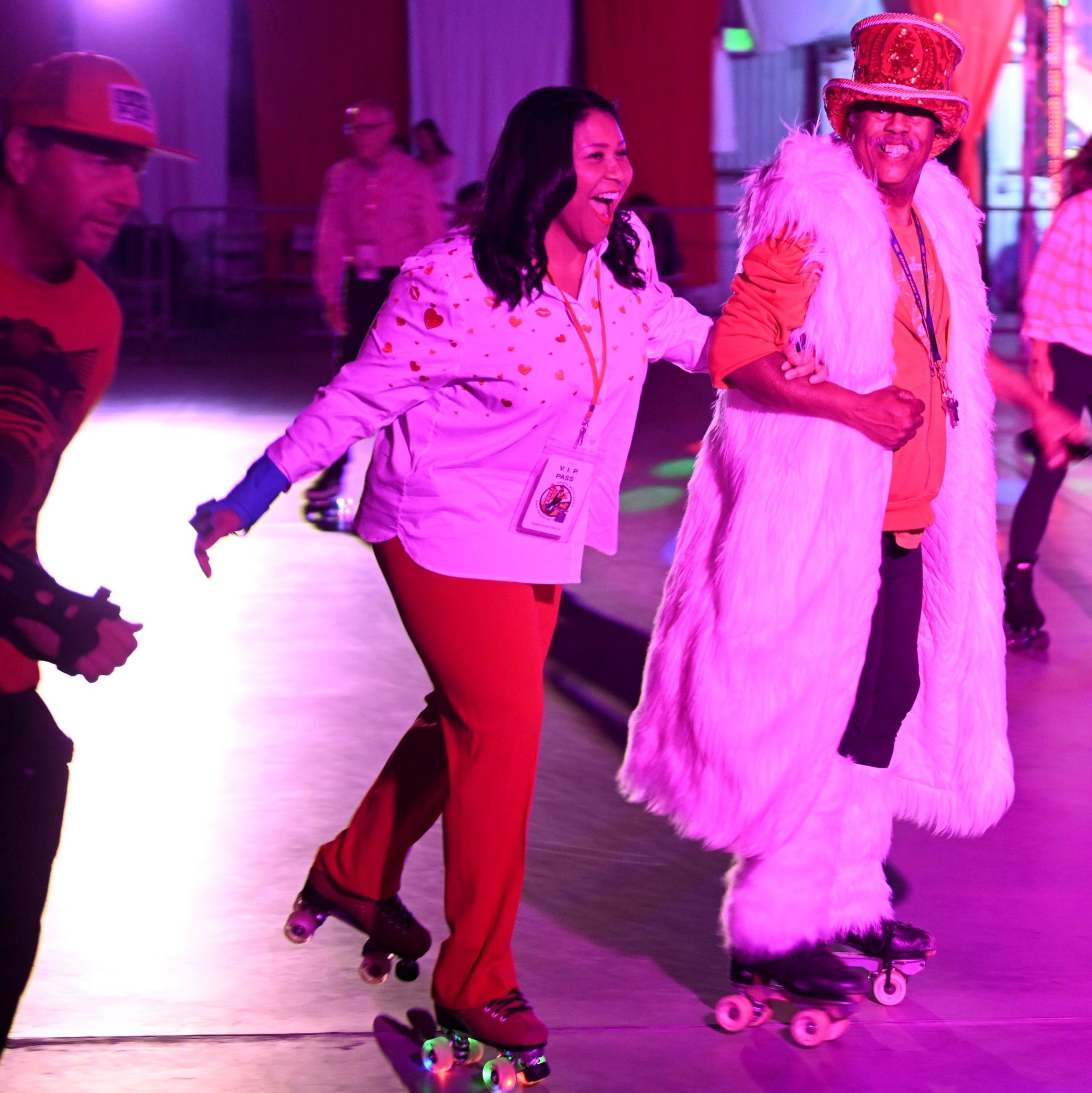 A group of people are roller skating indoors under vibrant colored lights. A woman in a white shirt and red pants smiles while skating beside a man in a fluffy outfit and top hat.