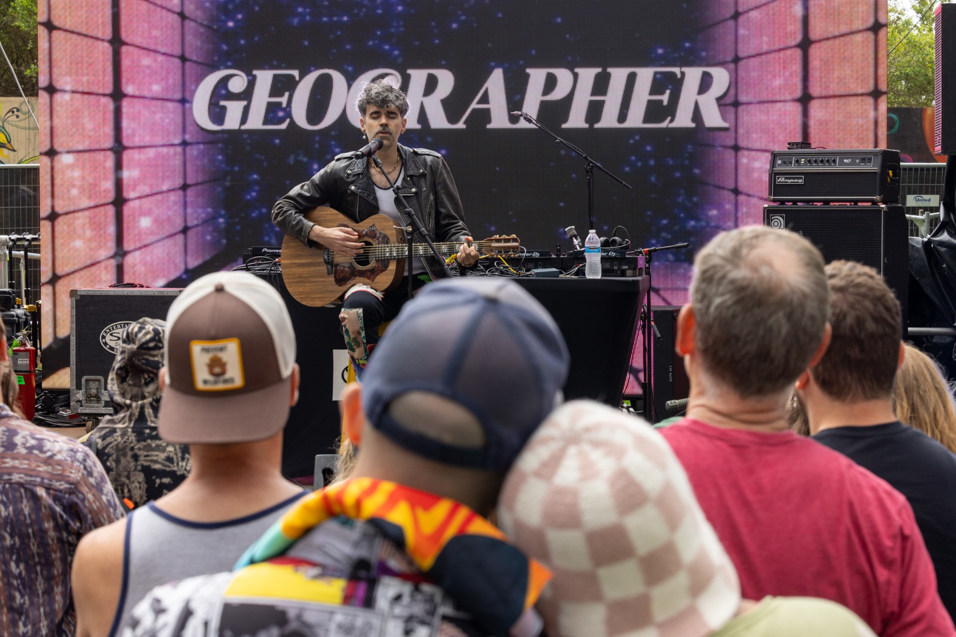 A musician in a leather jacket plays acoustic guitar and sings on stage while a small crowd watches, with "GEOGRAPHER" displayed behind him.
