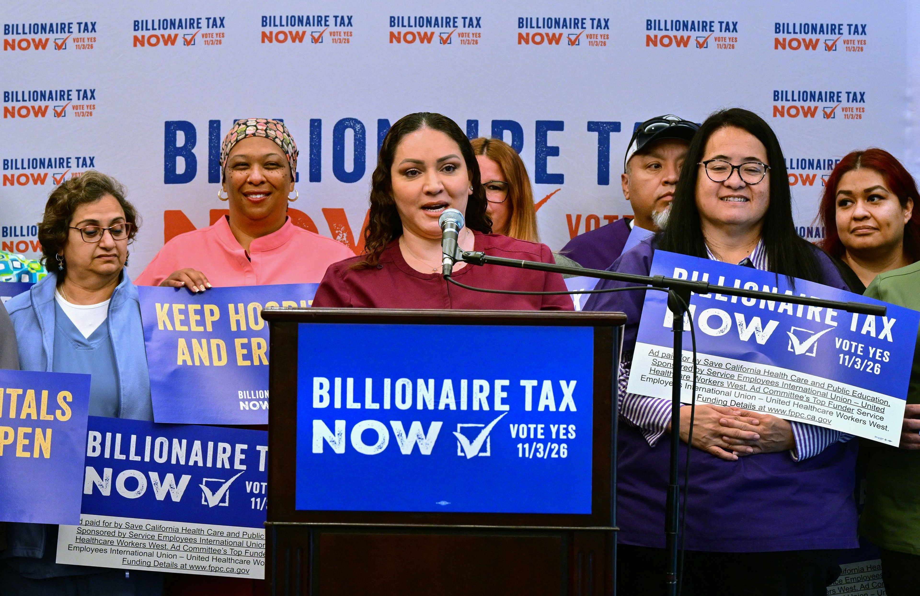 A diverse group of people stands behind a speaker at a podium with a sign reading “Billionaire Tax Now, Vote Yes 11/3/26,” holding supportive protest signs.