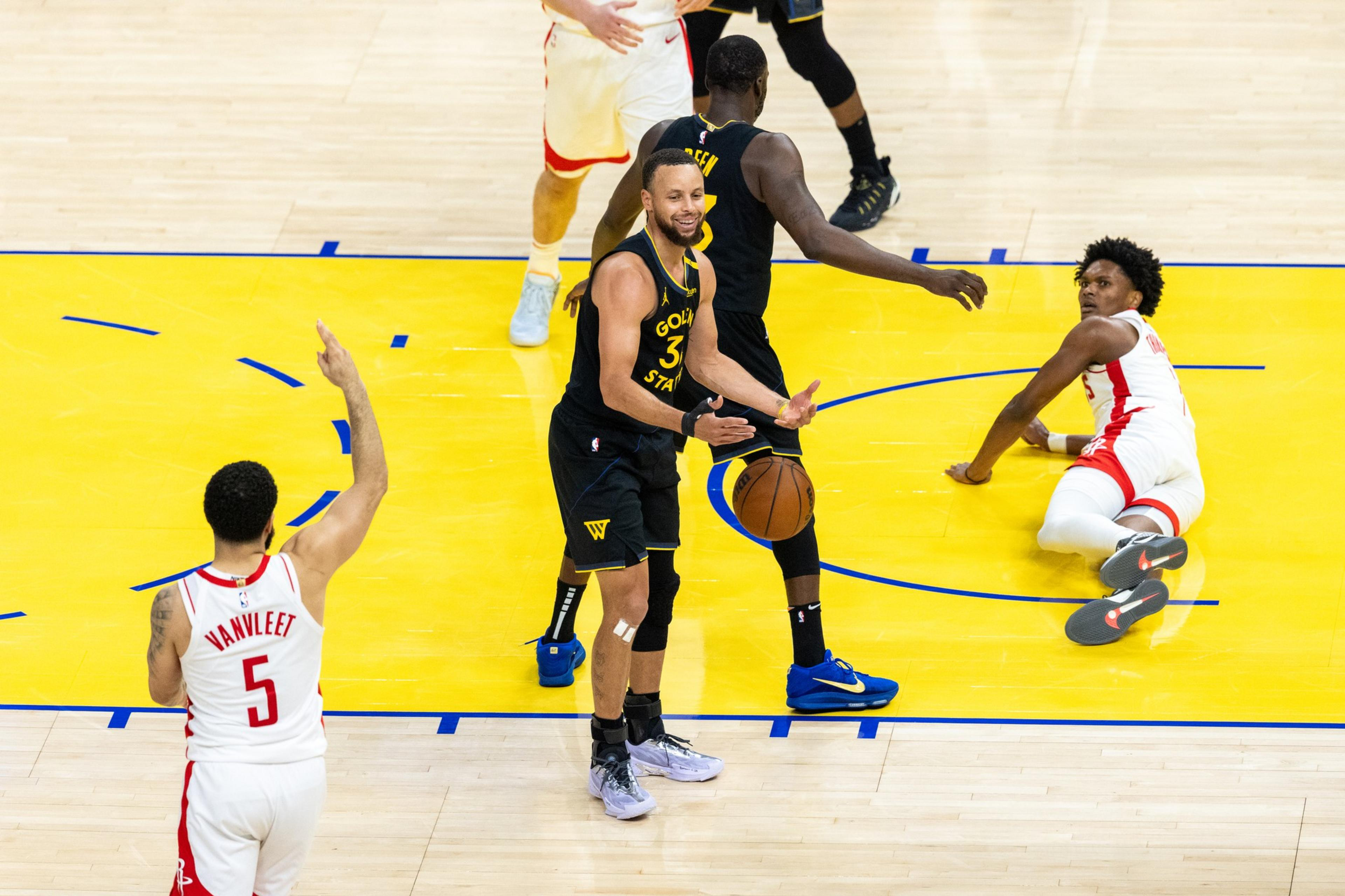 A basketball player in a black uniform is smiling and holding a ball, surrounded by players in red and black jerseys on a brightly lit court.